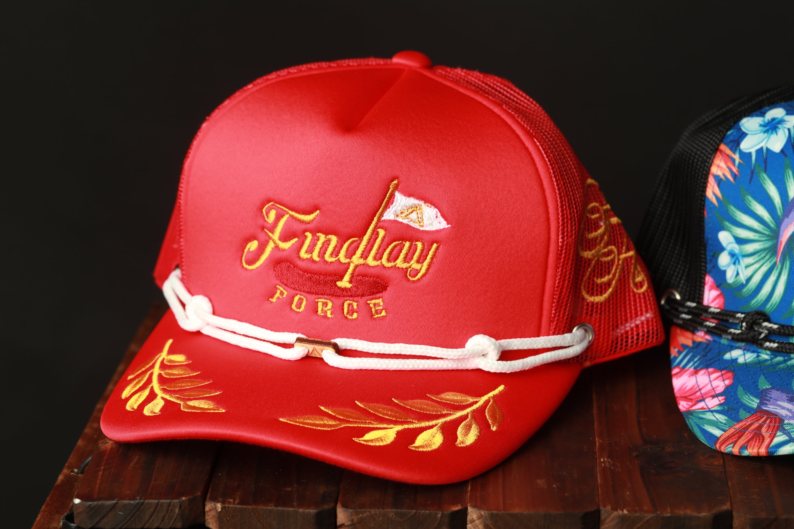 A red baseball cap with the text "Findlay FORCE" printed on it, sitting on a wooden surface next to another colorful cap.