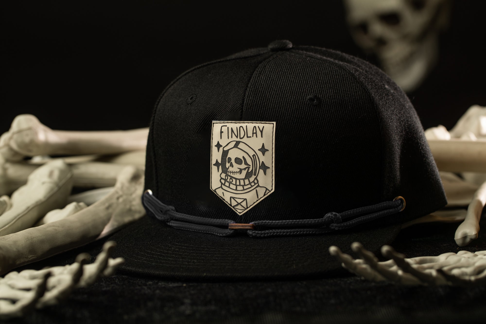 A black baseball cap with a skull and crossbones design and the word "FINDLAY" printed on it, placed on a surface with skeletal bones.
