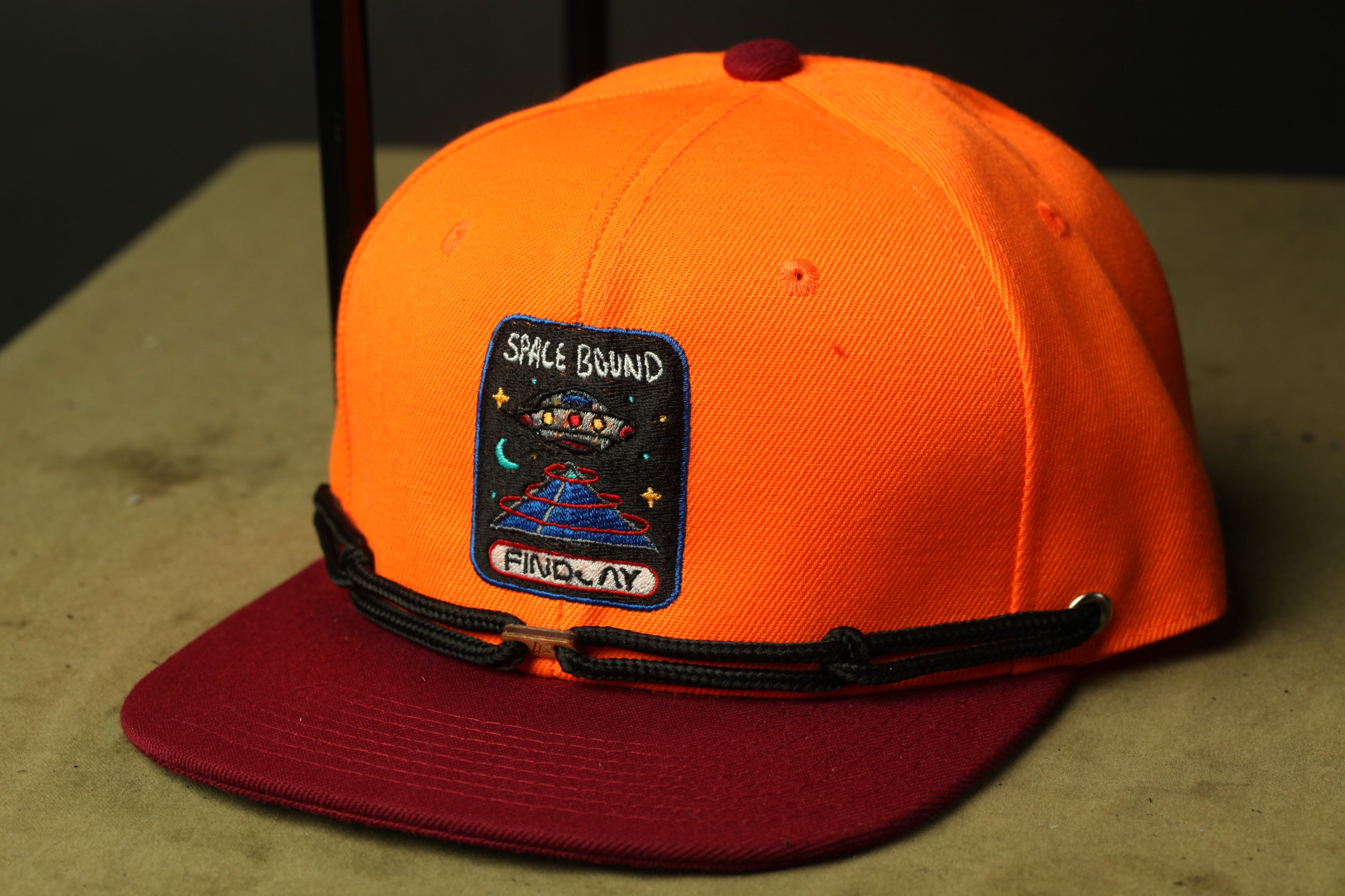 An orange baseball cap with a space-themed patch on it, sitting on a wooden surface.
