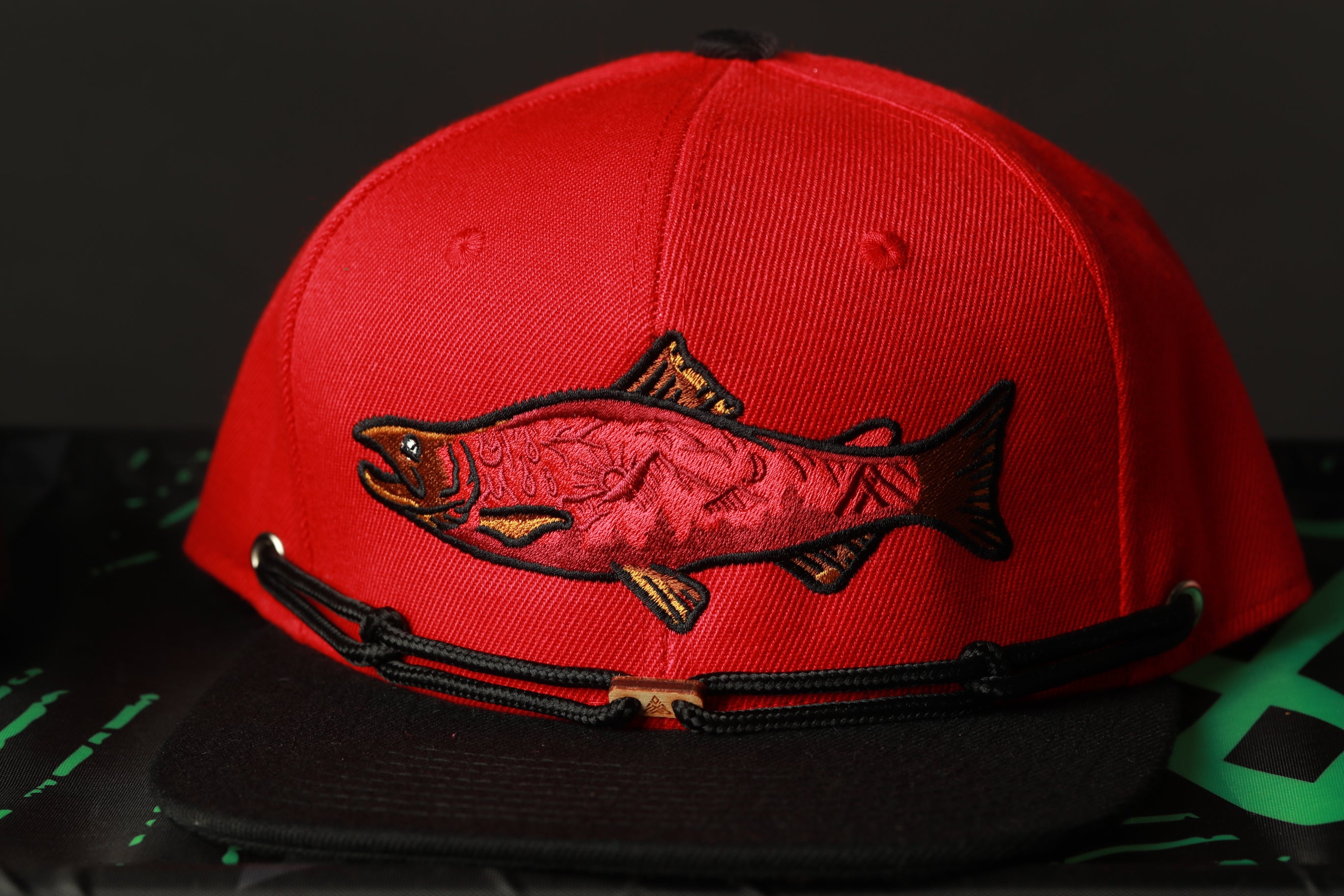 A red baseball cap with a fish embroidered on it, placed on a black surface.