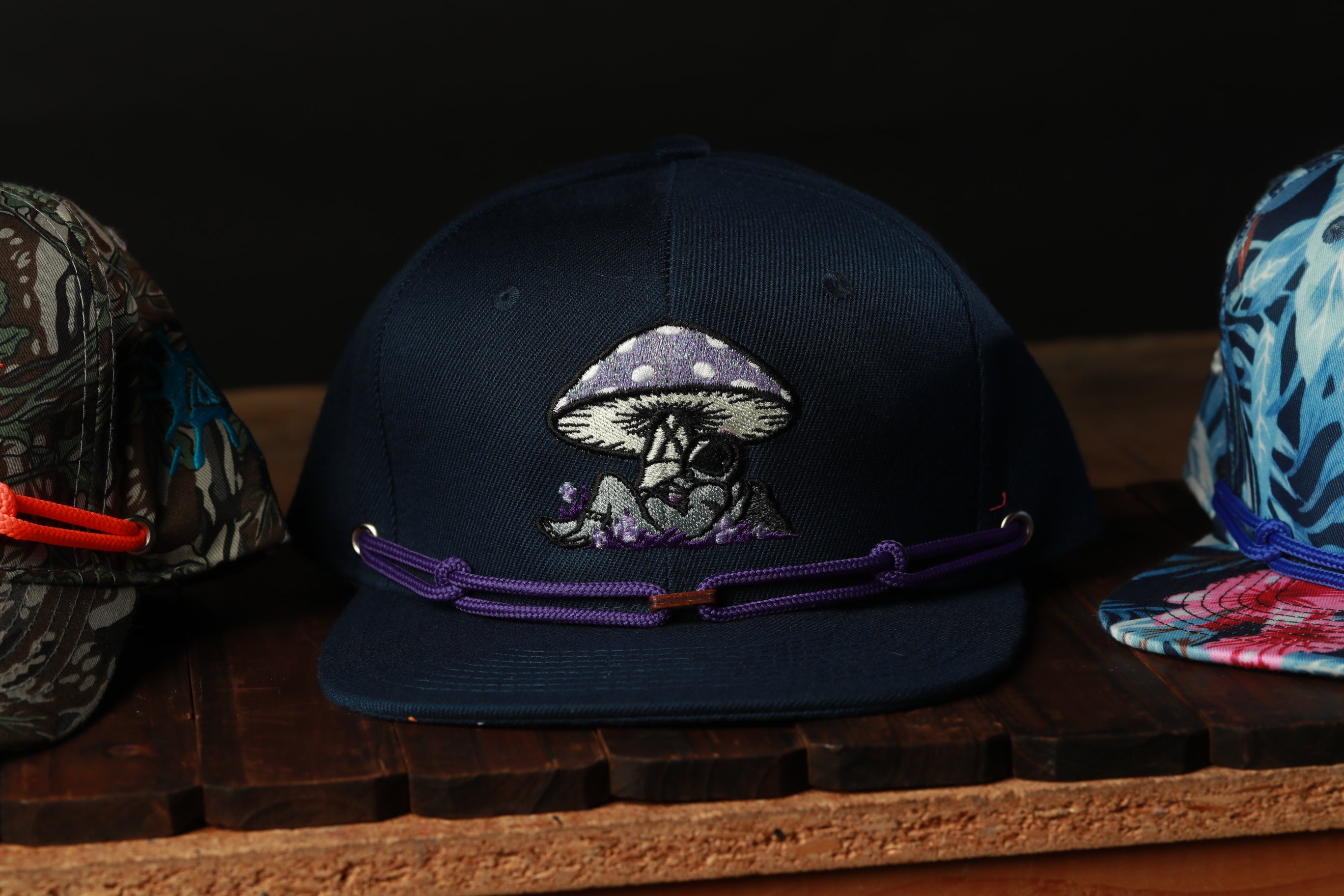 A dark blue baseball cap with a mushroom design and purple straps is displayed on a wooden surface.