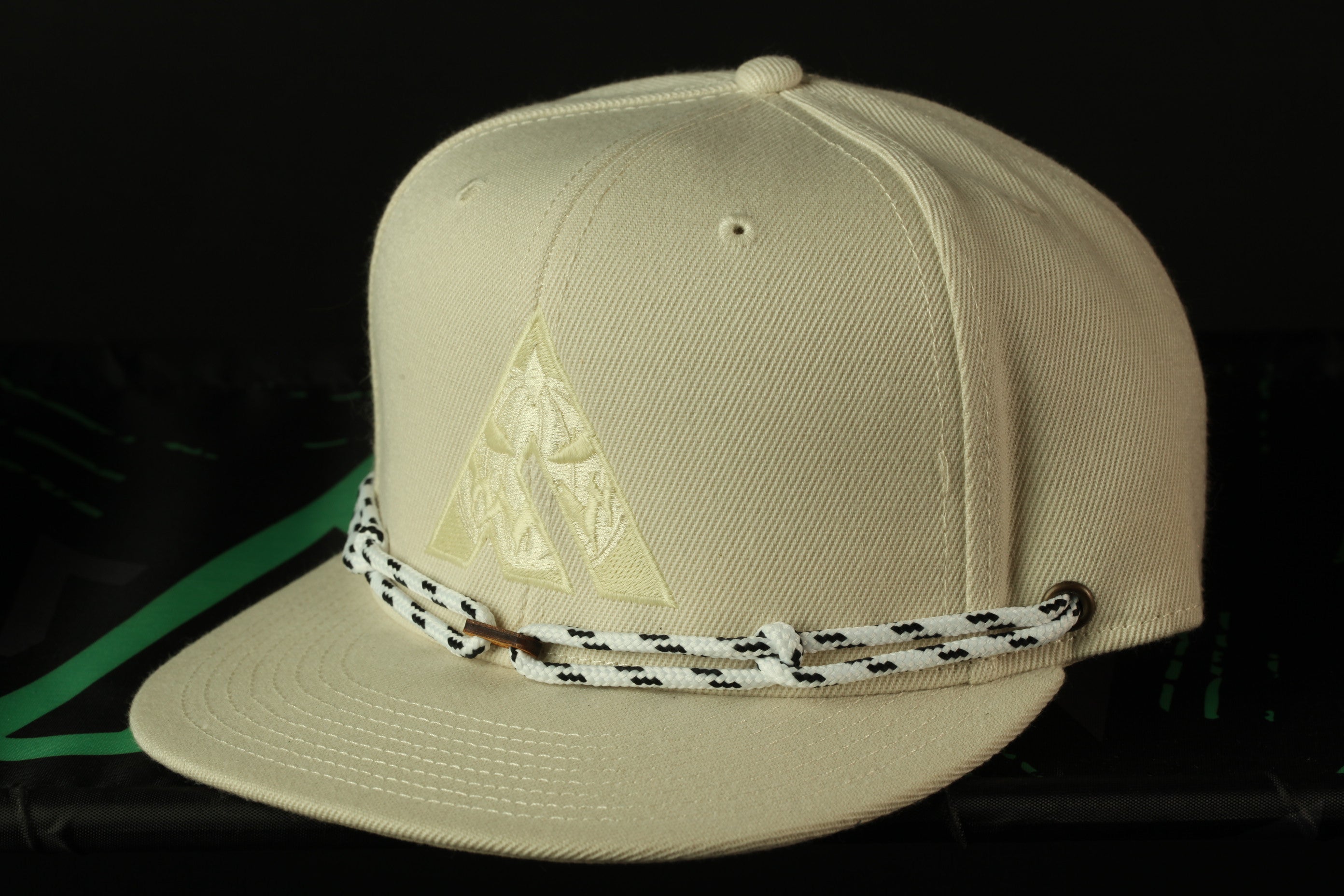 A beige baseball cap with a white and black braided strap is shown against a black background.
