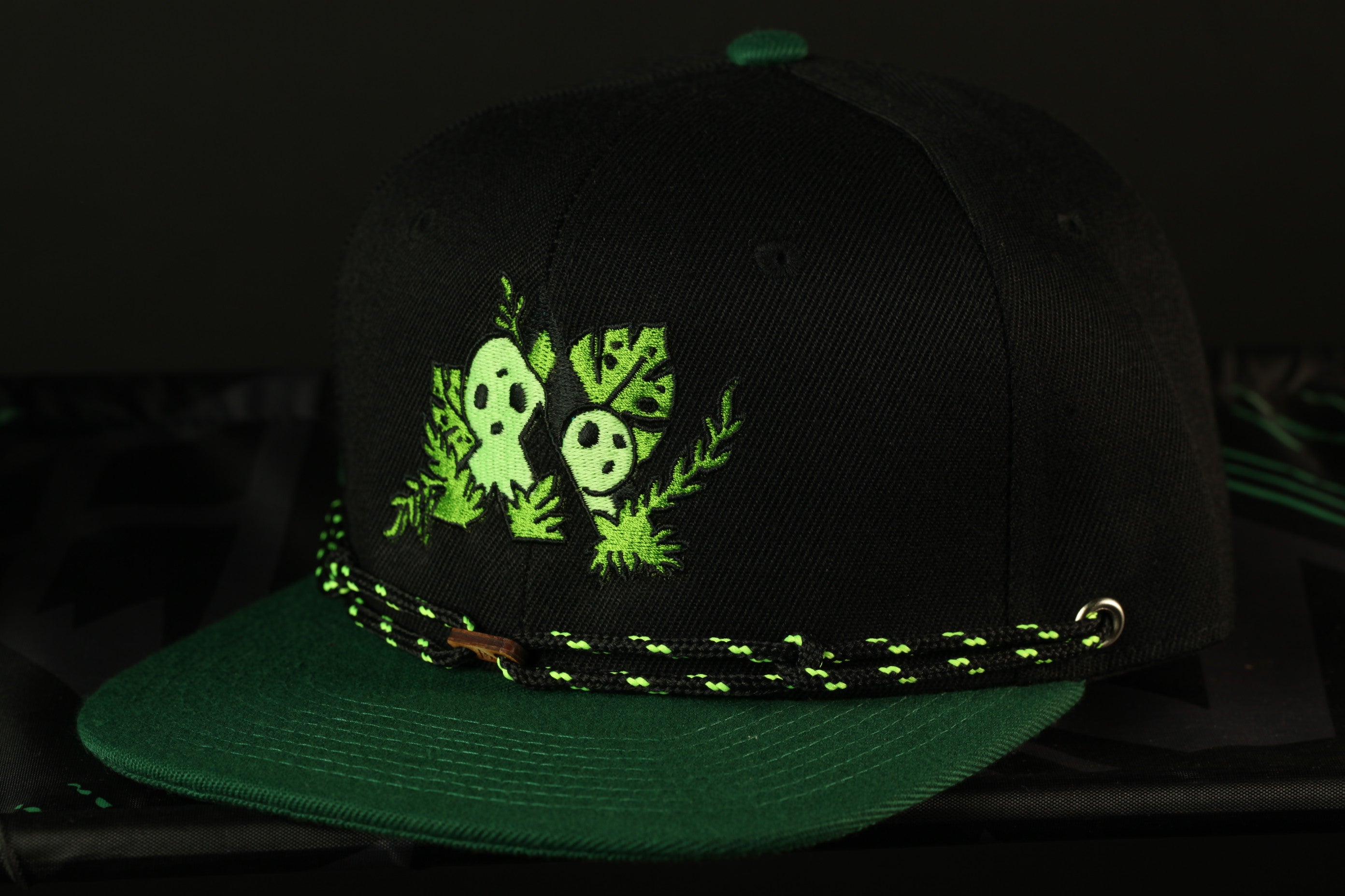 A black baseball cap with a green and black braided band, featuring a green and white design with two cartoon-like faces and leaves.