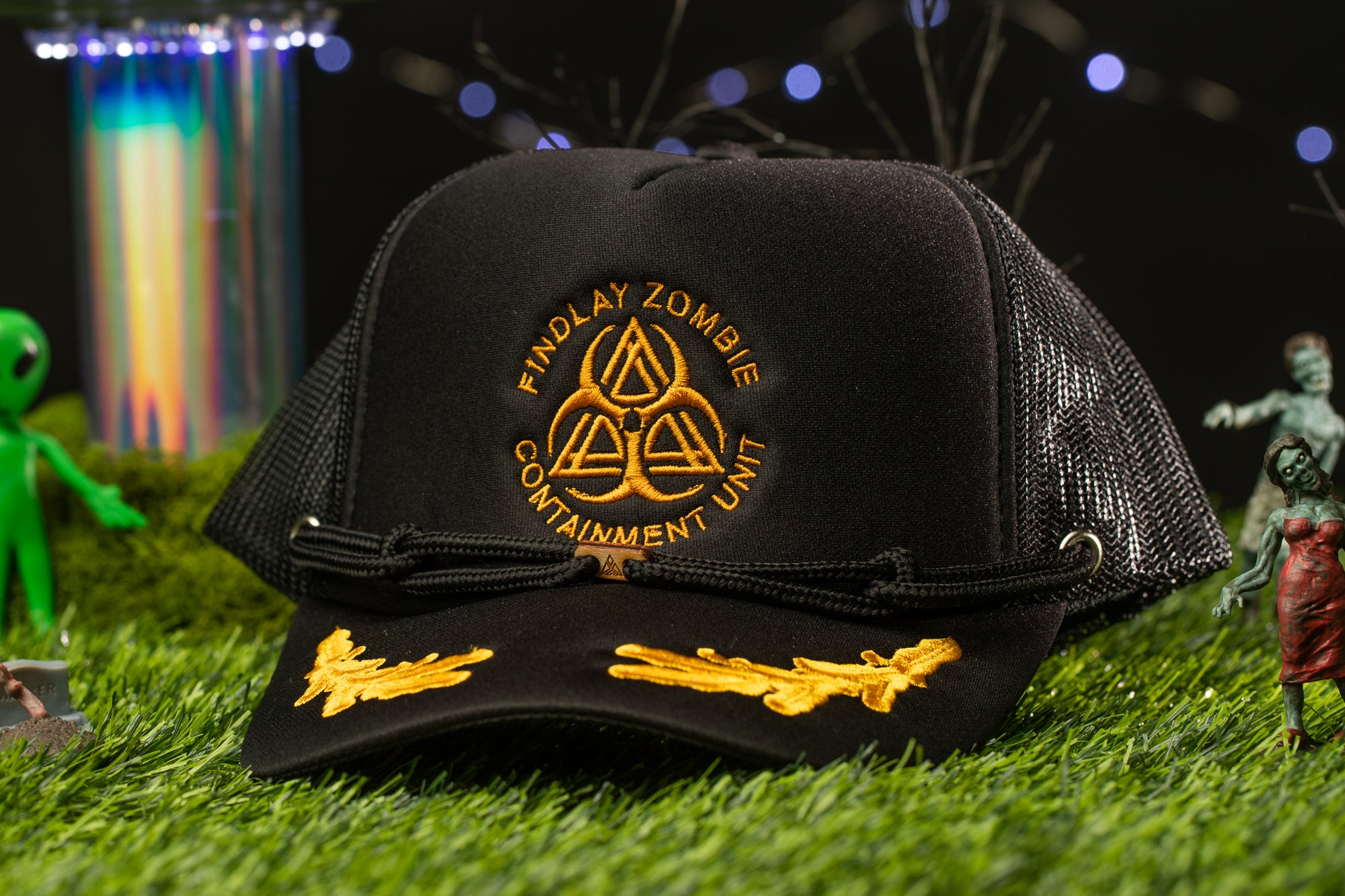 A black baseball cap with gold embroidered designs and text, placed on a grassy surface with various small figurines and a colorful, blurred background.