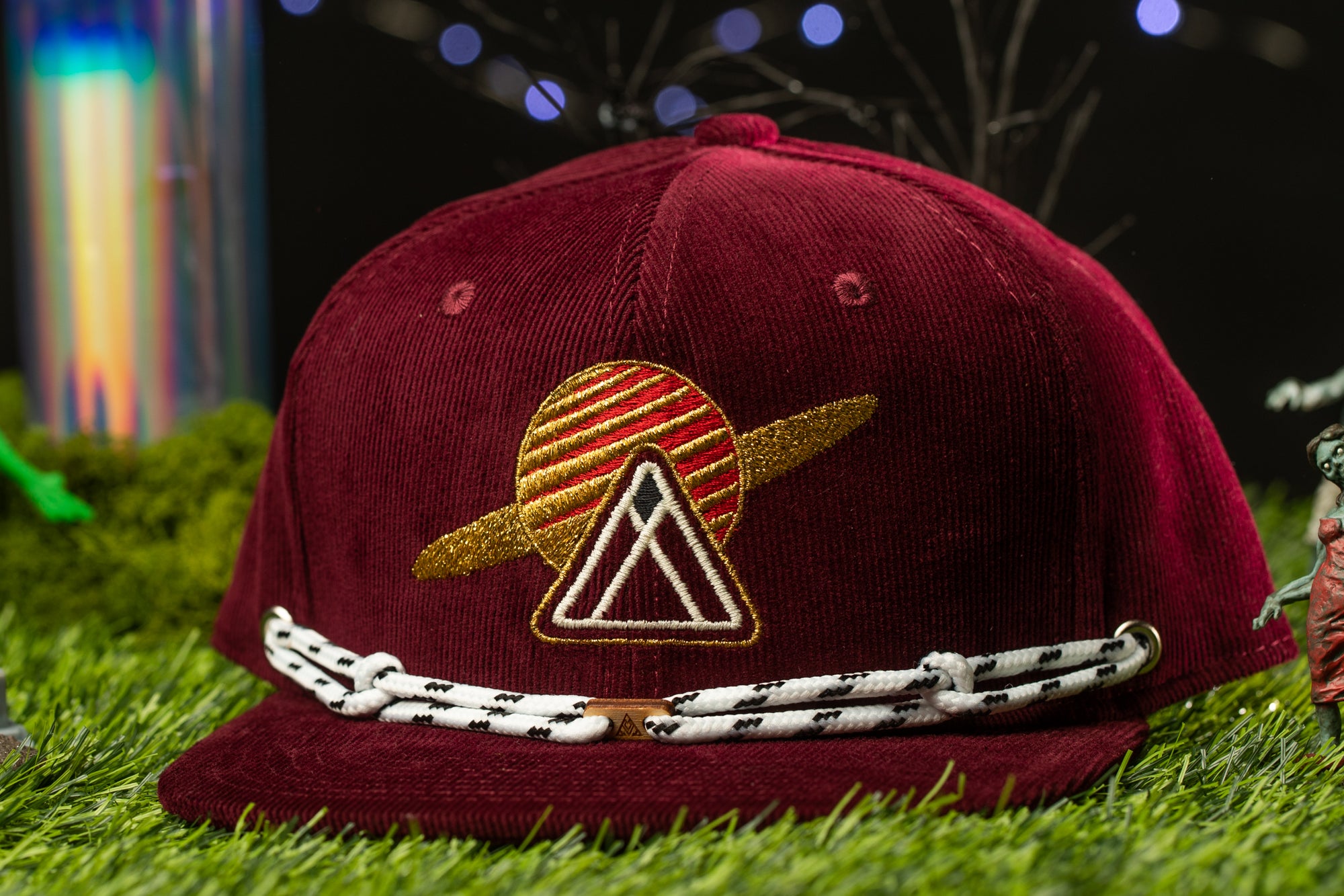 A burgundy baseball cap with a gold and red striped design and a triangular emblem on the front, placed on a grassy surface with a blurred background.