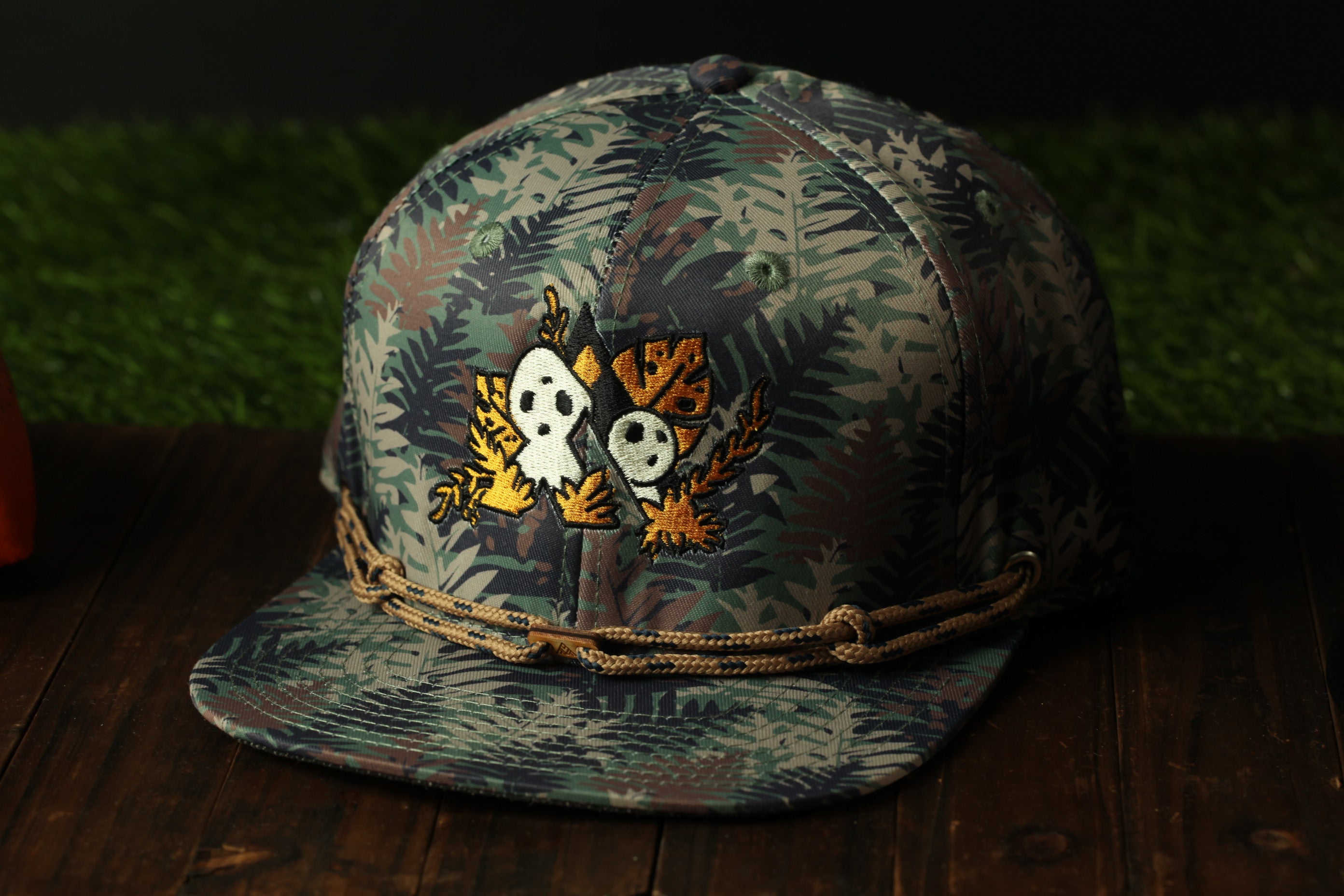 A camouflage baseball cap with a skull and leaf design on the front, sitting on a wooden surface.