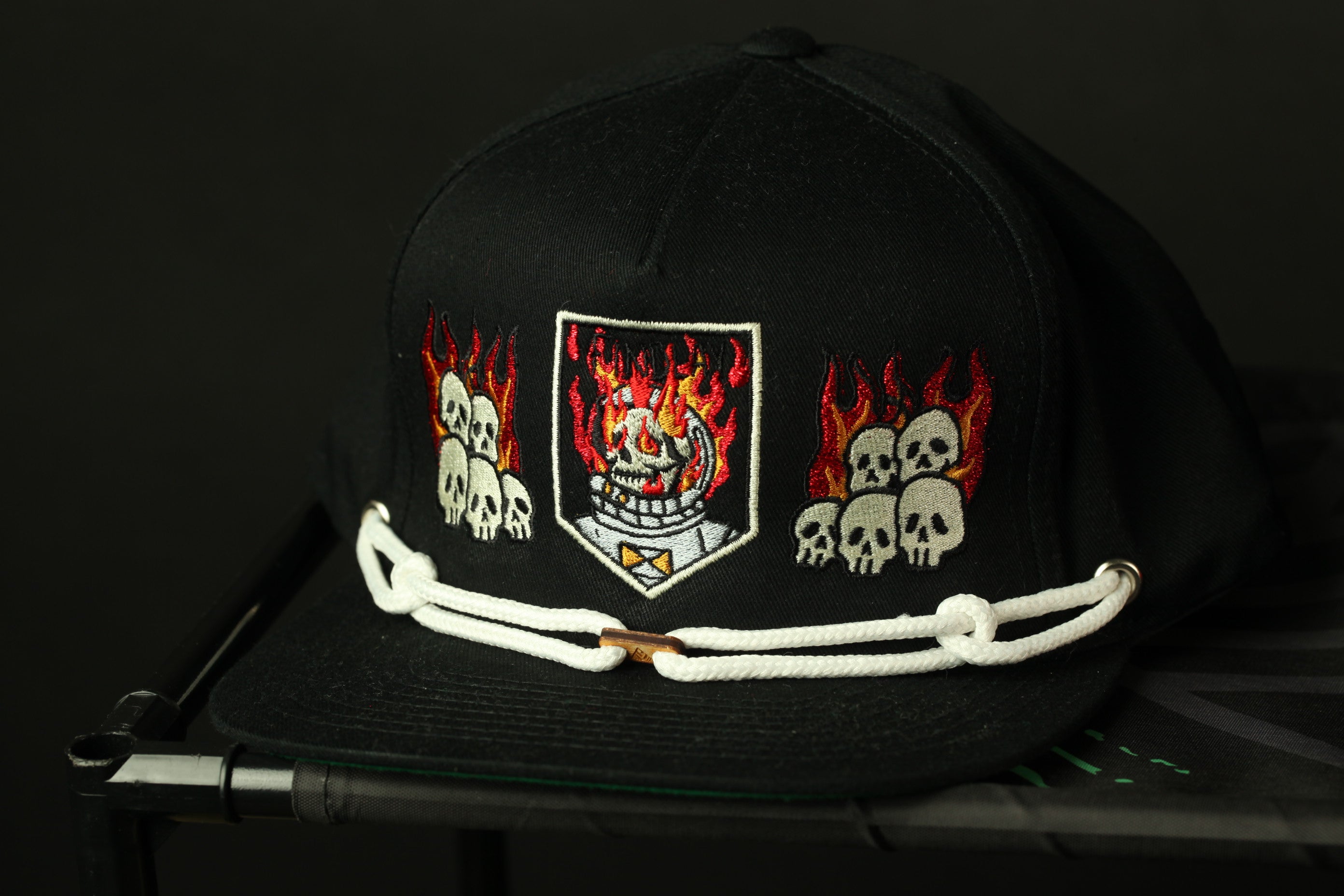 A black baseball cap with a white band and a central emblem featuring a skull and flames.