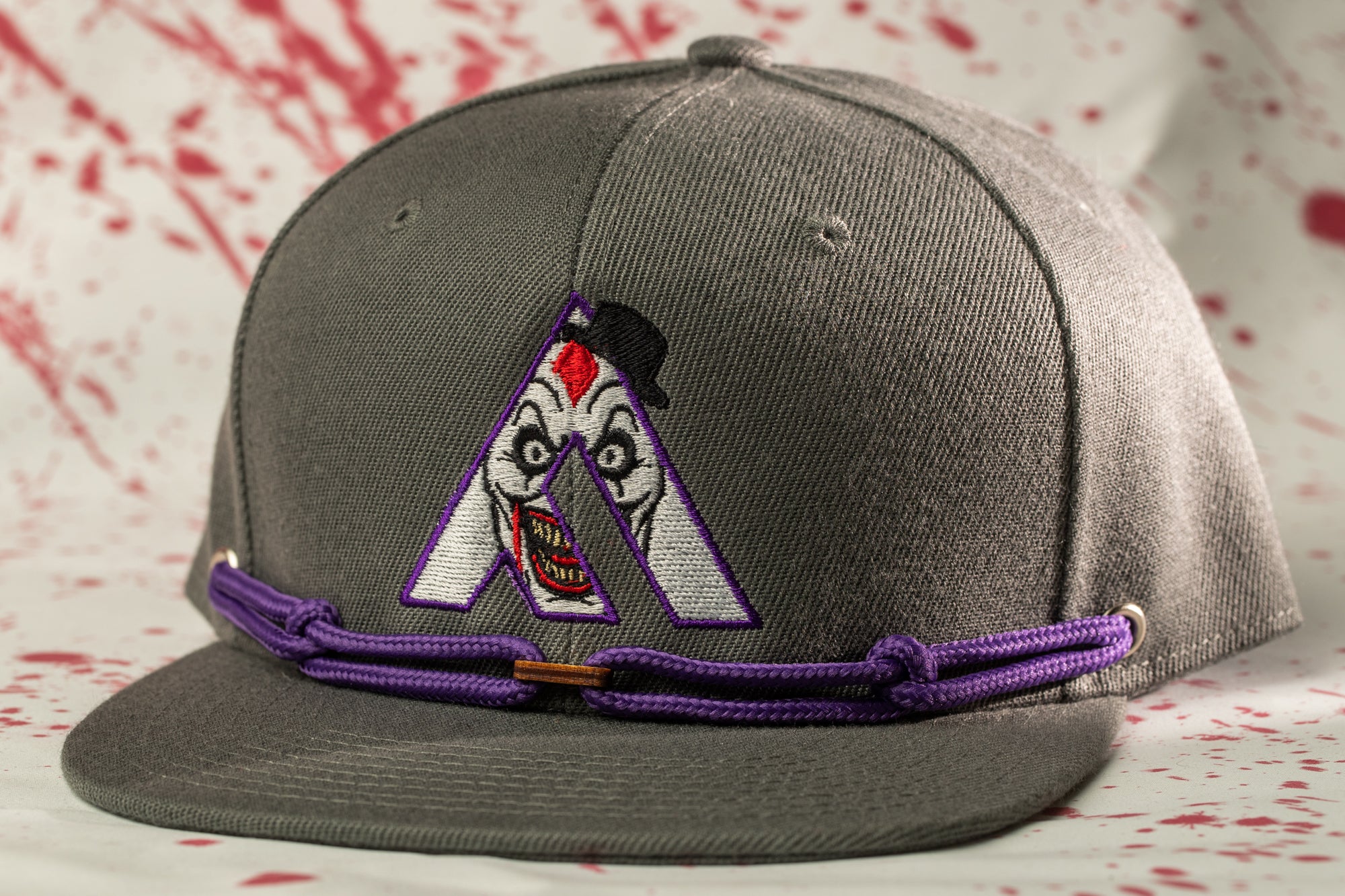 A gray baseball cap with a purple string and a purple and white clown face embroidered on the front.