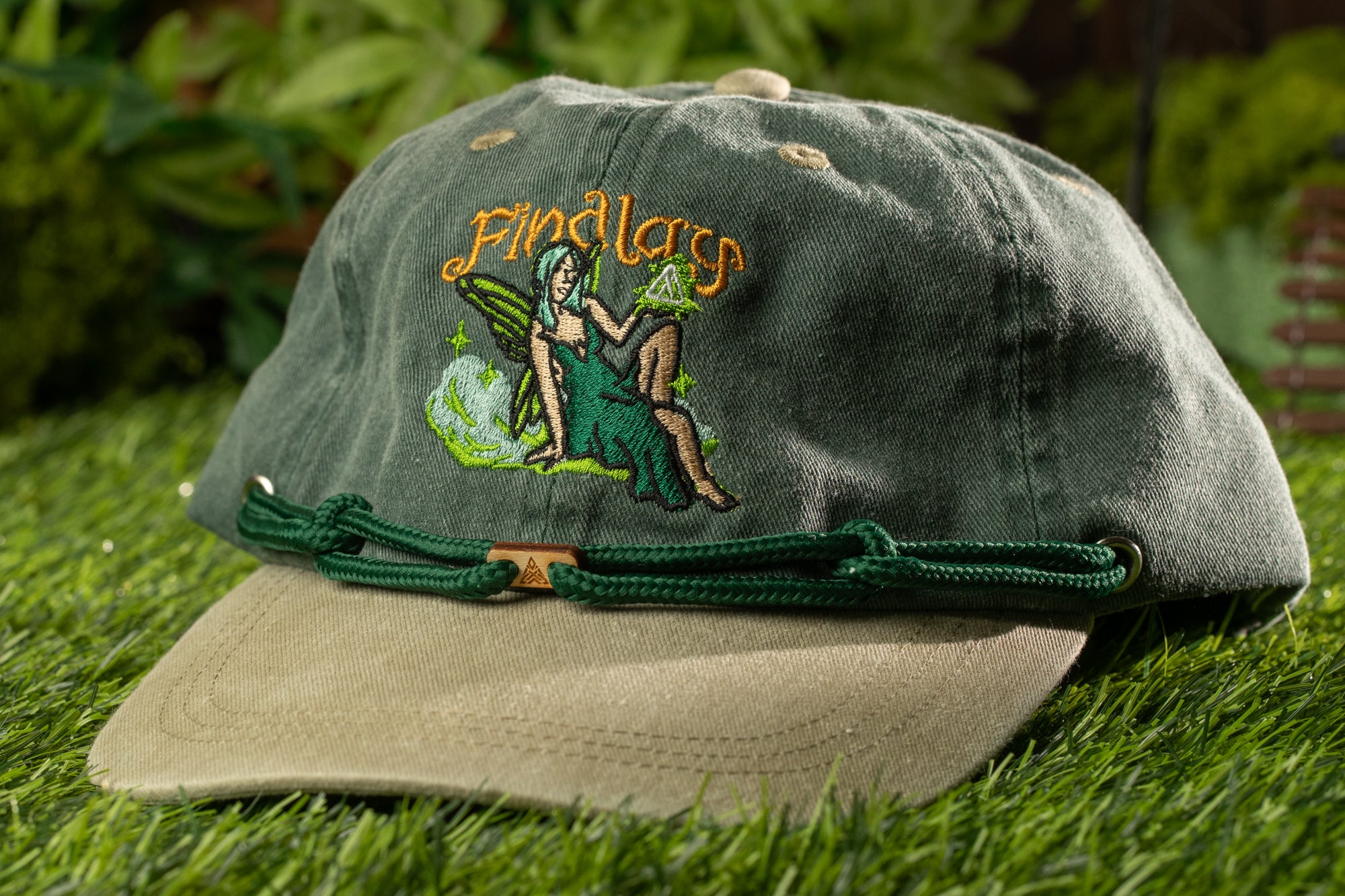 A green baseball cap with a green strap and a small embroidered image of a woman on the front, sitting on a grassy surface.