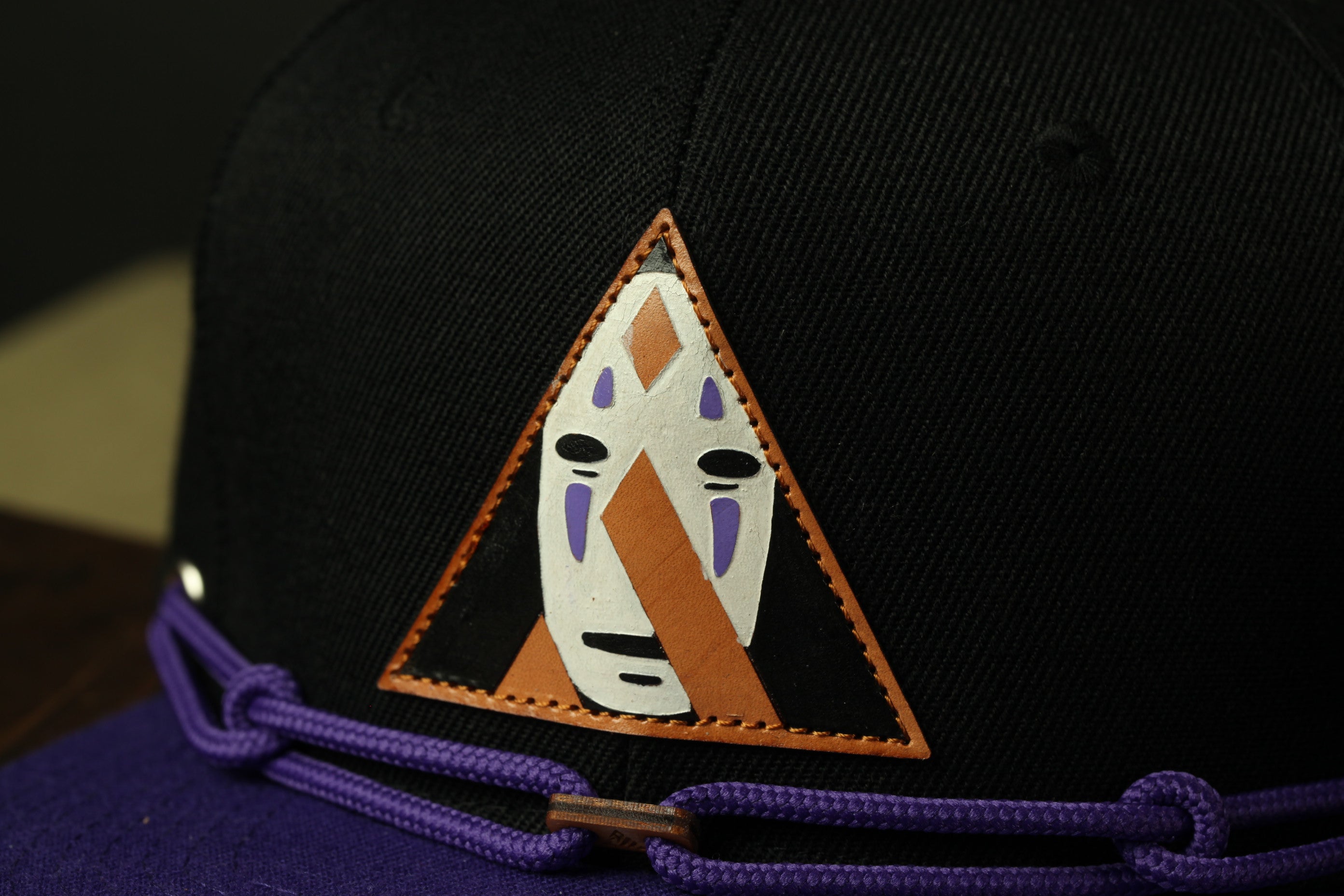 A close-up view of a black baseball cap with a unique triangular logo featuring a face design.