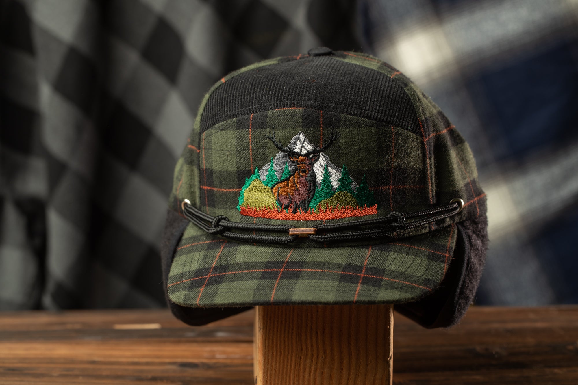 A plaid hat with a mountain and animal embroidered on it, displayed on a wooden stand.