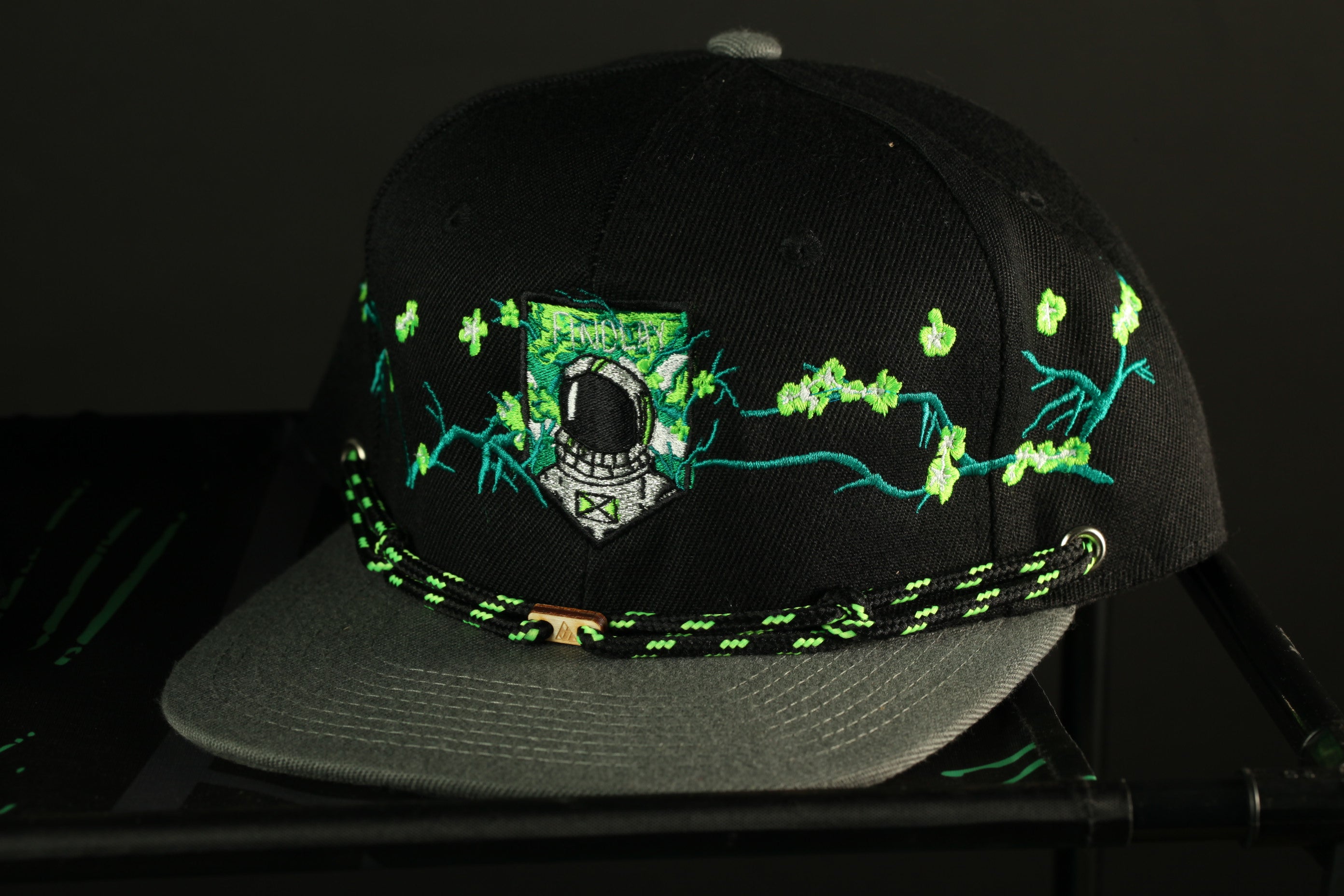 A black baseball cap with a green and white floral design and a small emblem on the front, displayed on a black surface.