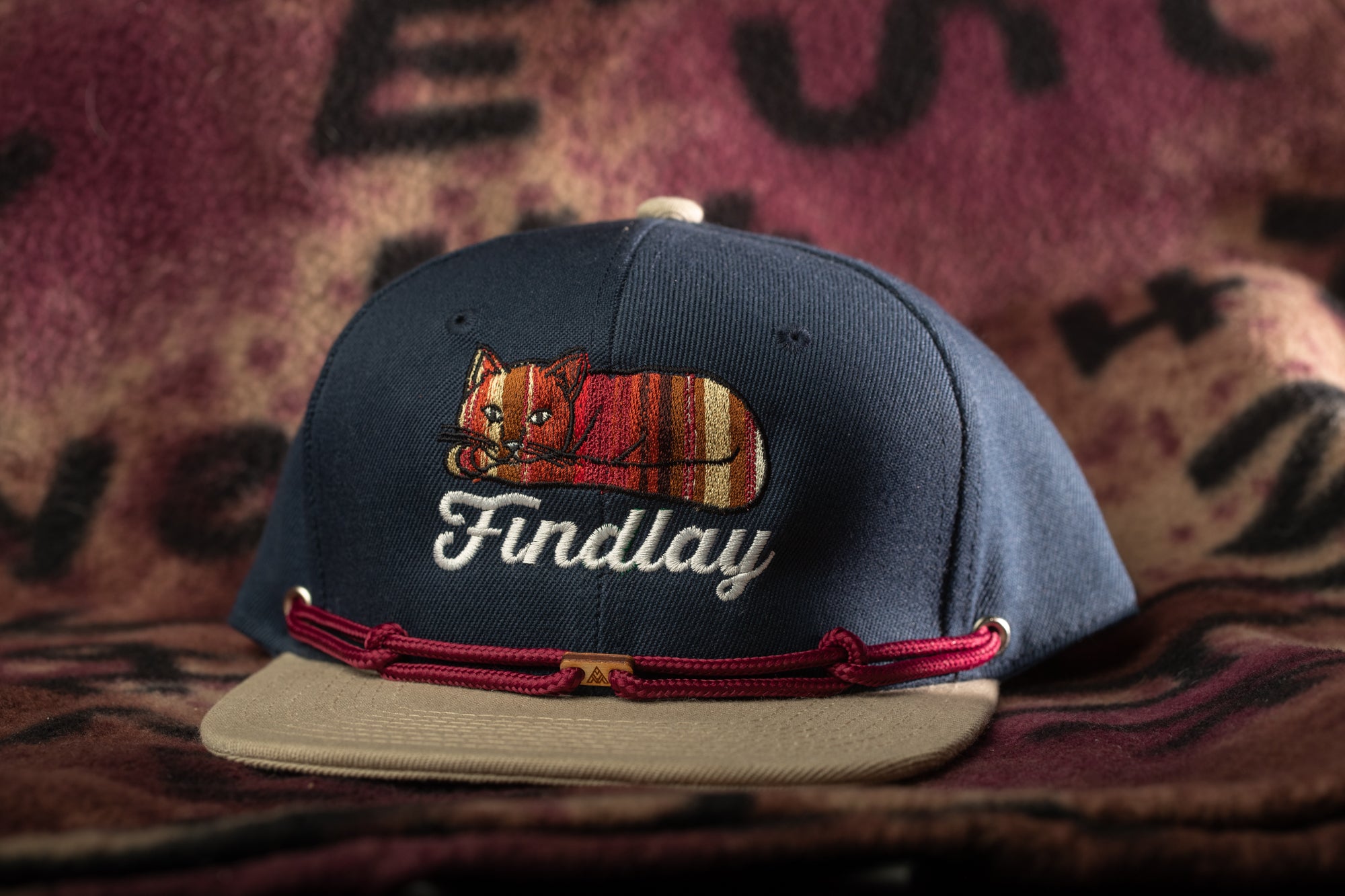 A navy blue baseball cap with a cat embroidered on it, sitting on a beige fabric surface.