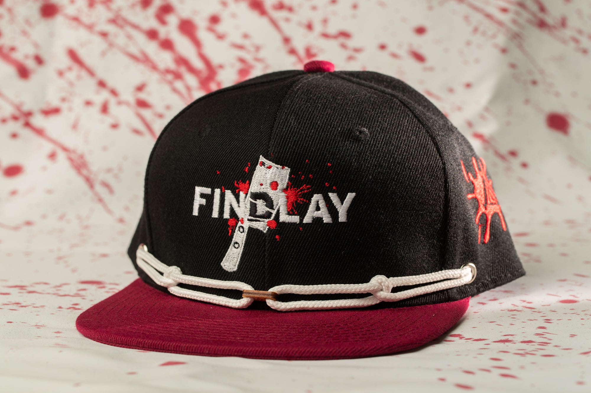 A black baseball cap with a red brim and a white chain strap, featuring a graphic of a knife and the word "FINDlay" printed on it.