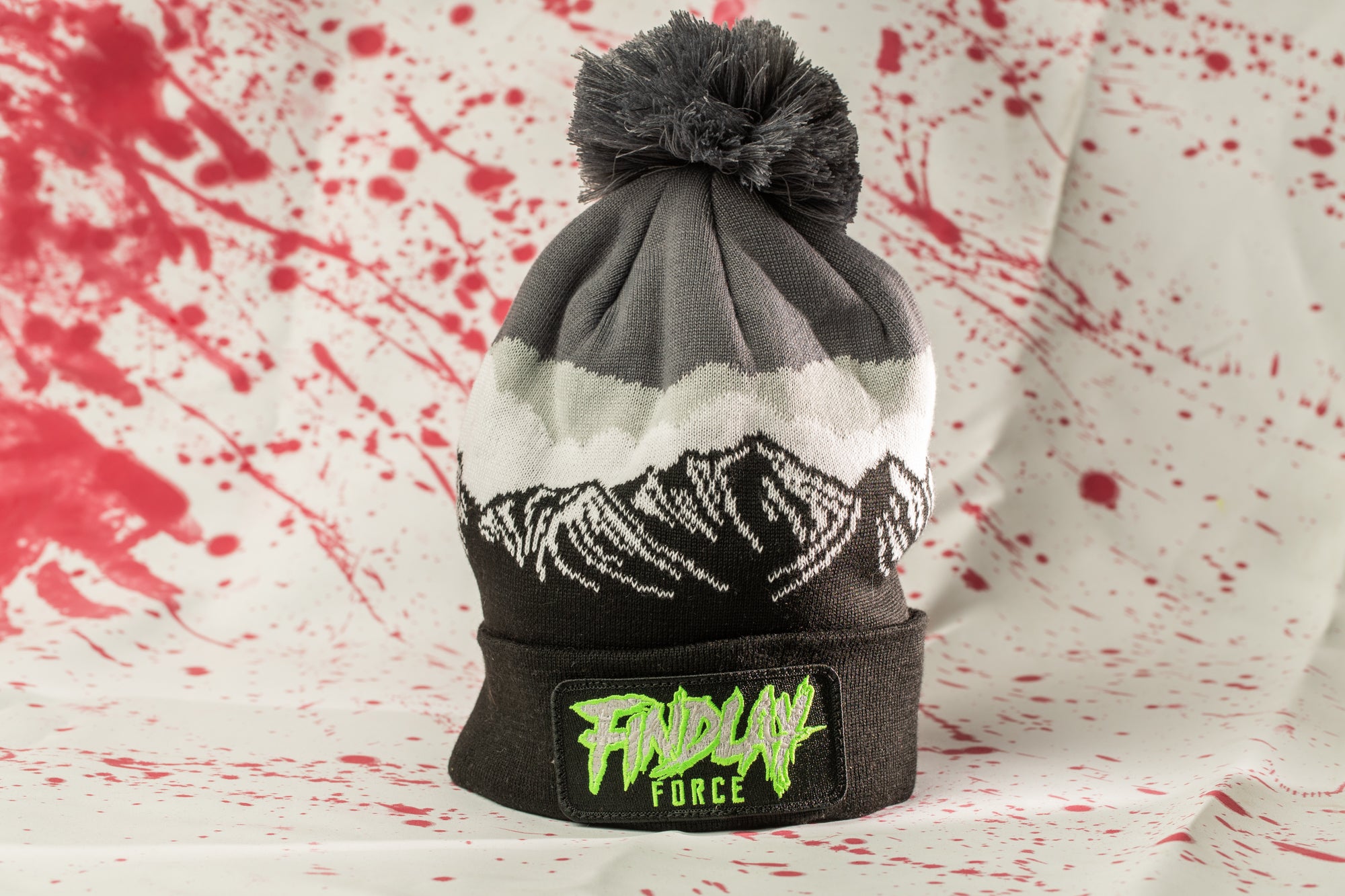 A black and gray beanie with a pom pom and a green patch on the front, featuring a graphic design, is displayed against a background with red splatters.