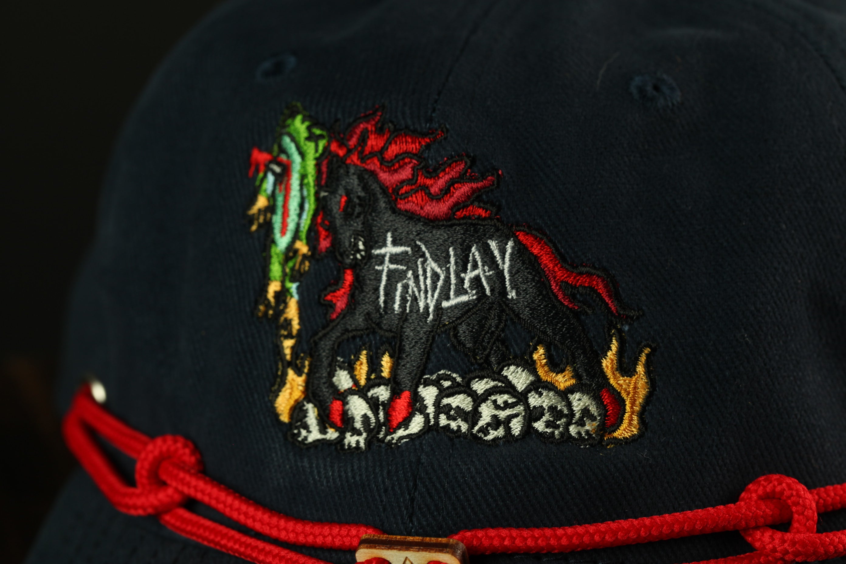 a close-up view of a black baseball cap with a red string around the brim. The cap has an embroidered design on the front, depicting a stylized horse with flames on its back.