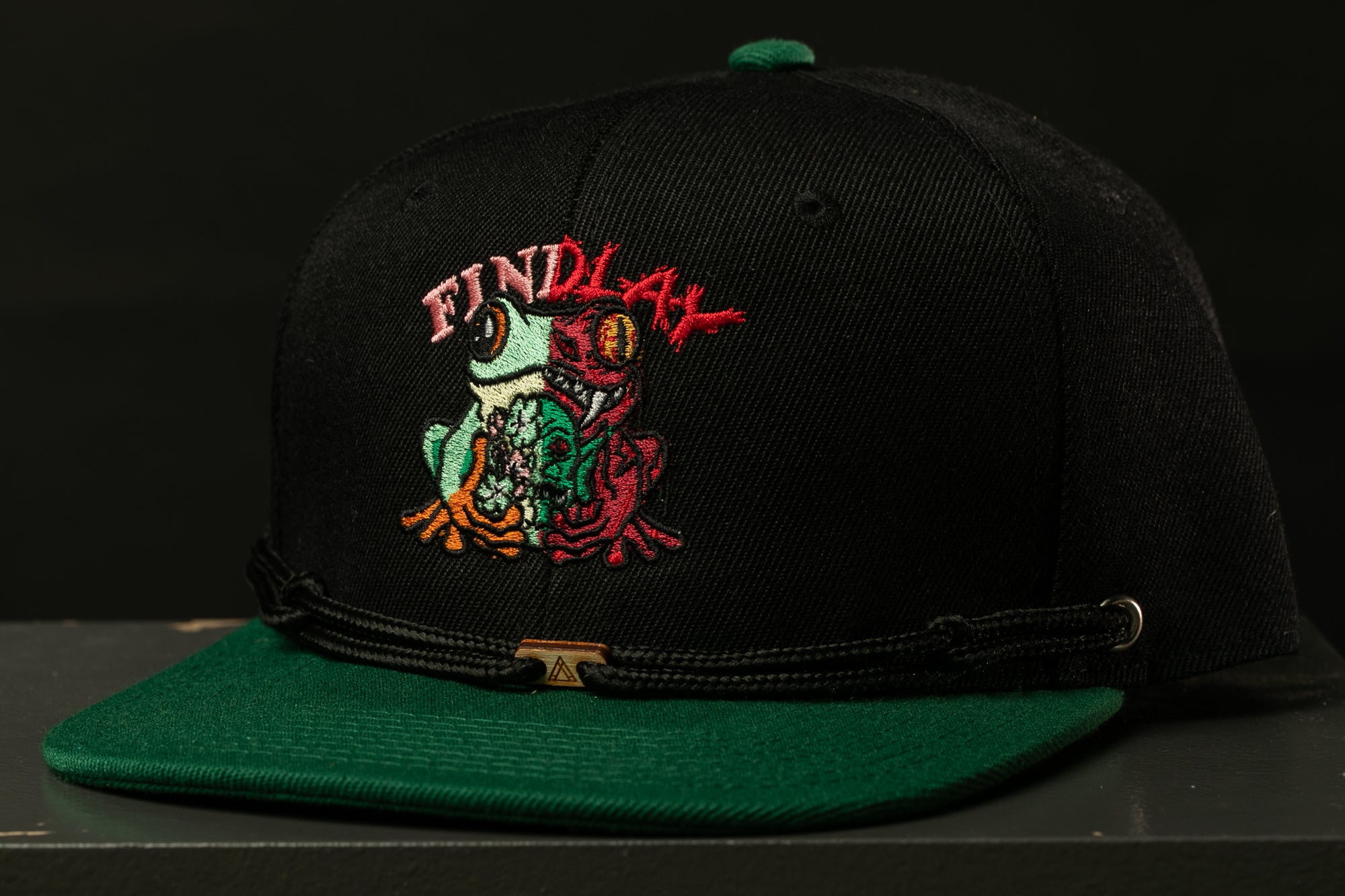 A black baseball cap with a green brim and a colorful frog embroidered on the front.
