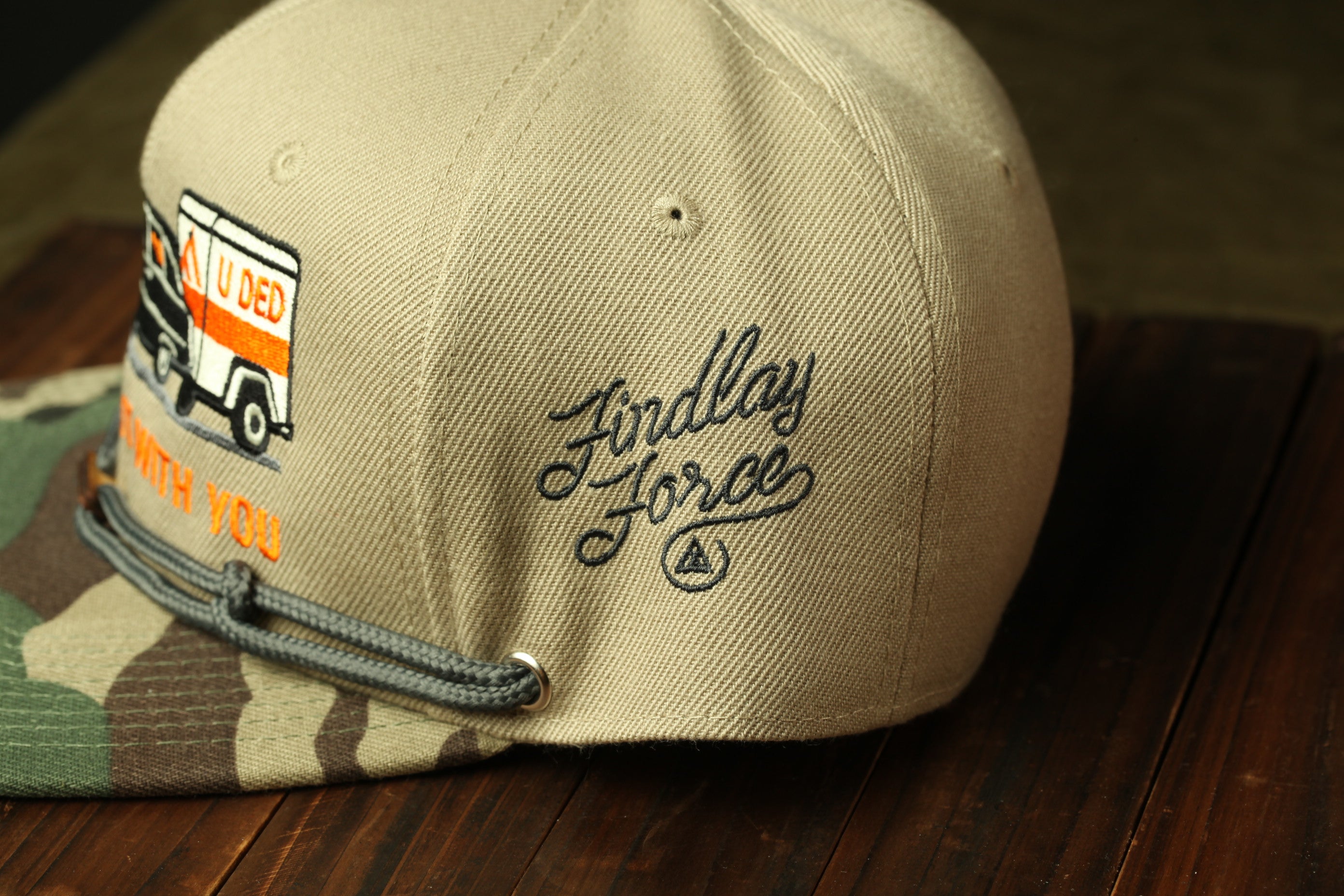 A beige baseball cap with a camouflage pattern and a logo on the side, placed on a wooden surface.