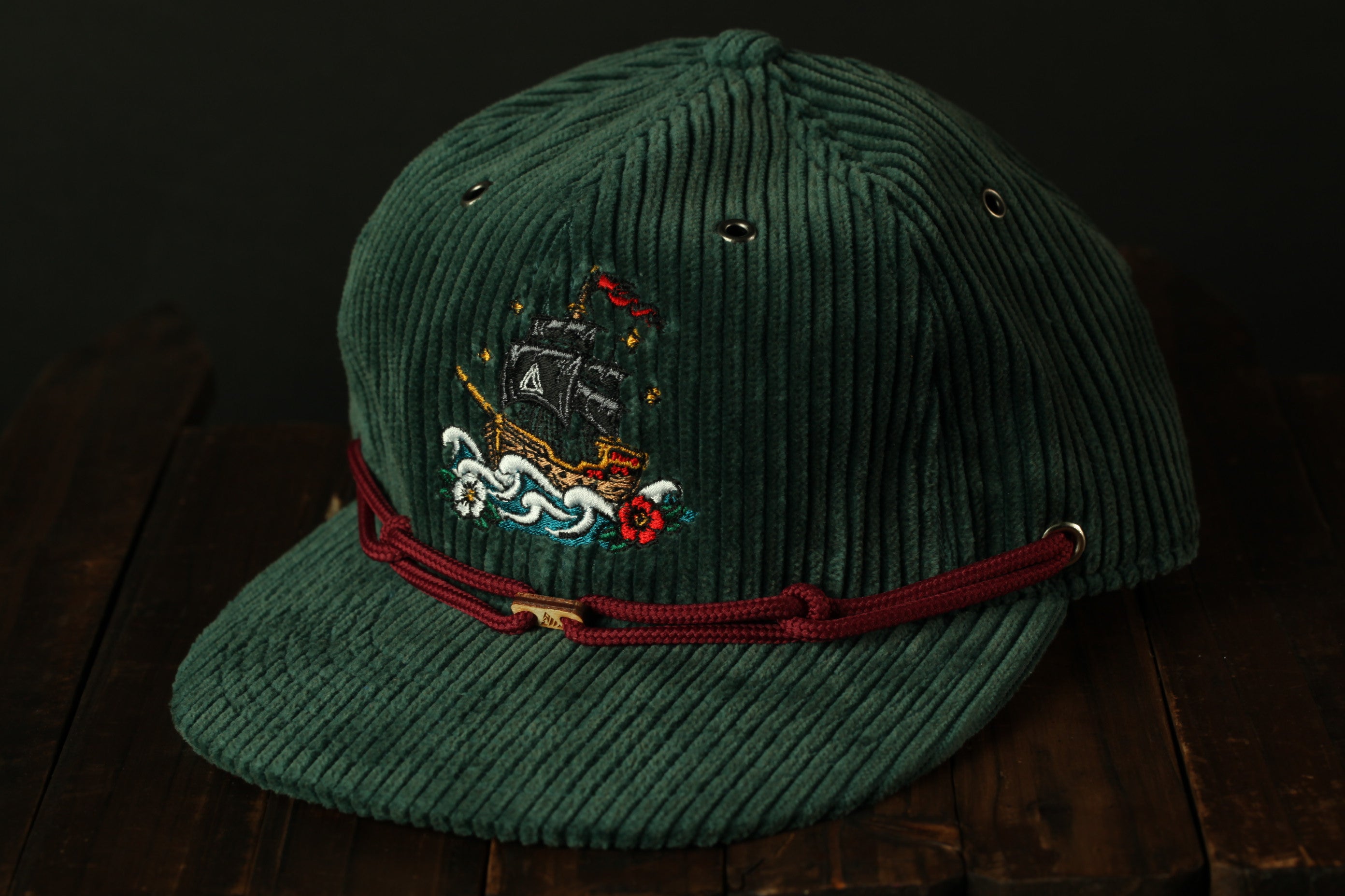 A green baseball cap with a red ribbon and a ship embroidered on it, placed on a wooden surface.
