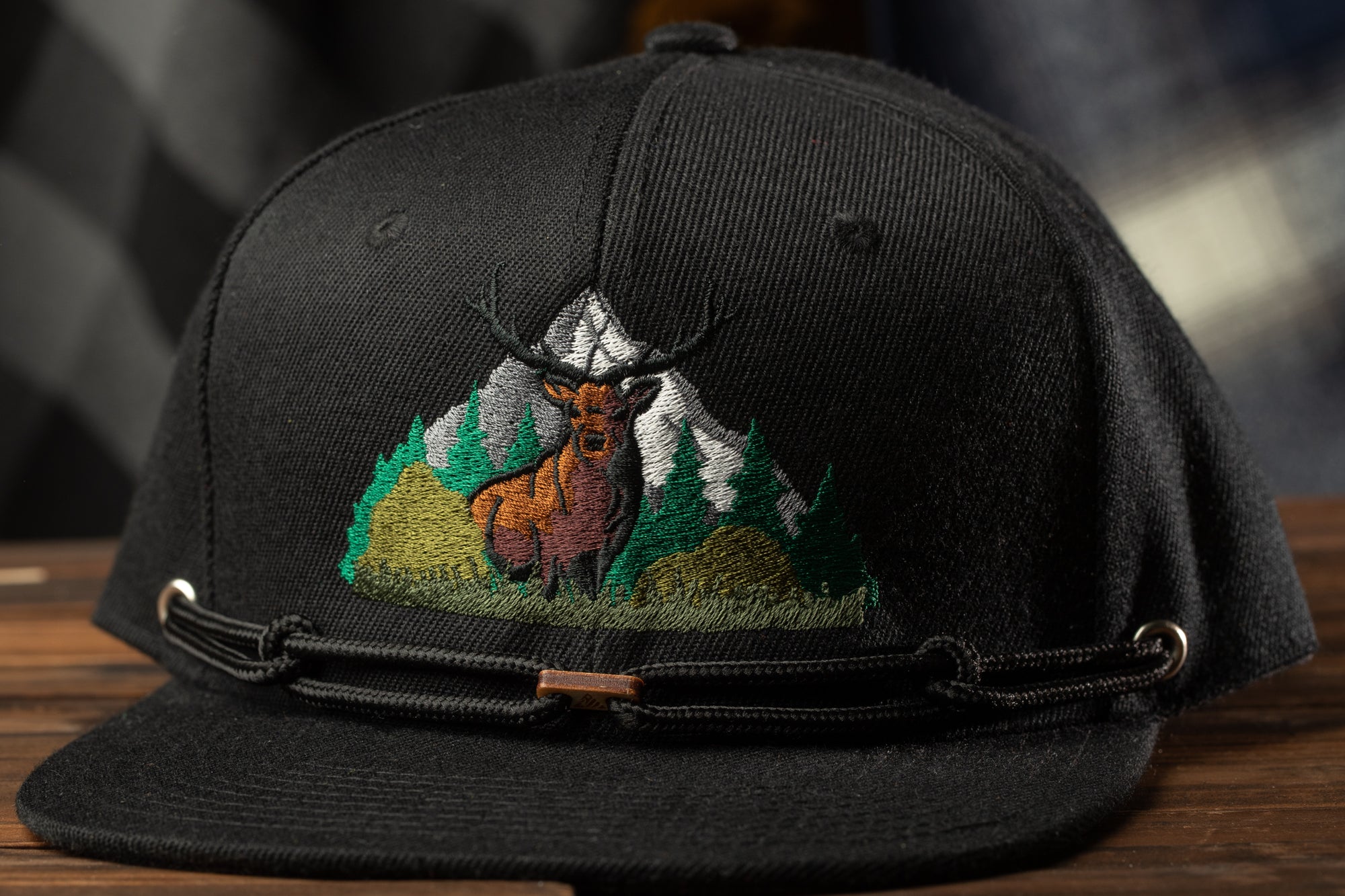 A black baseball cap with a detailed embroidered design depicting a deer standing in a mountainous landscape.