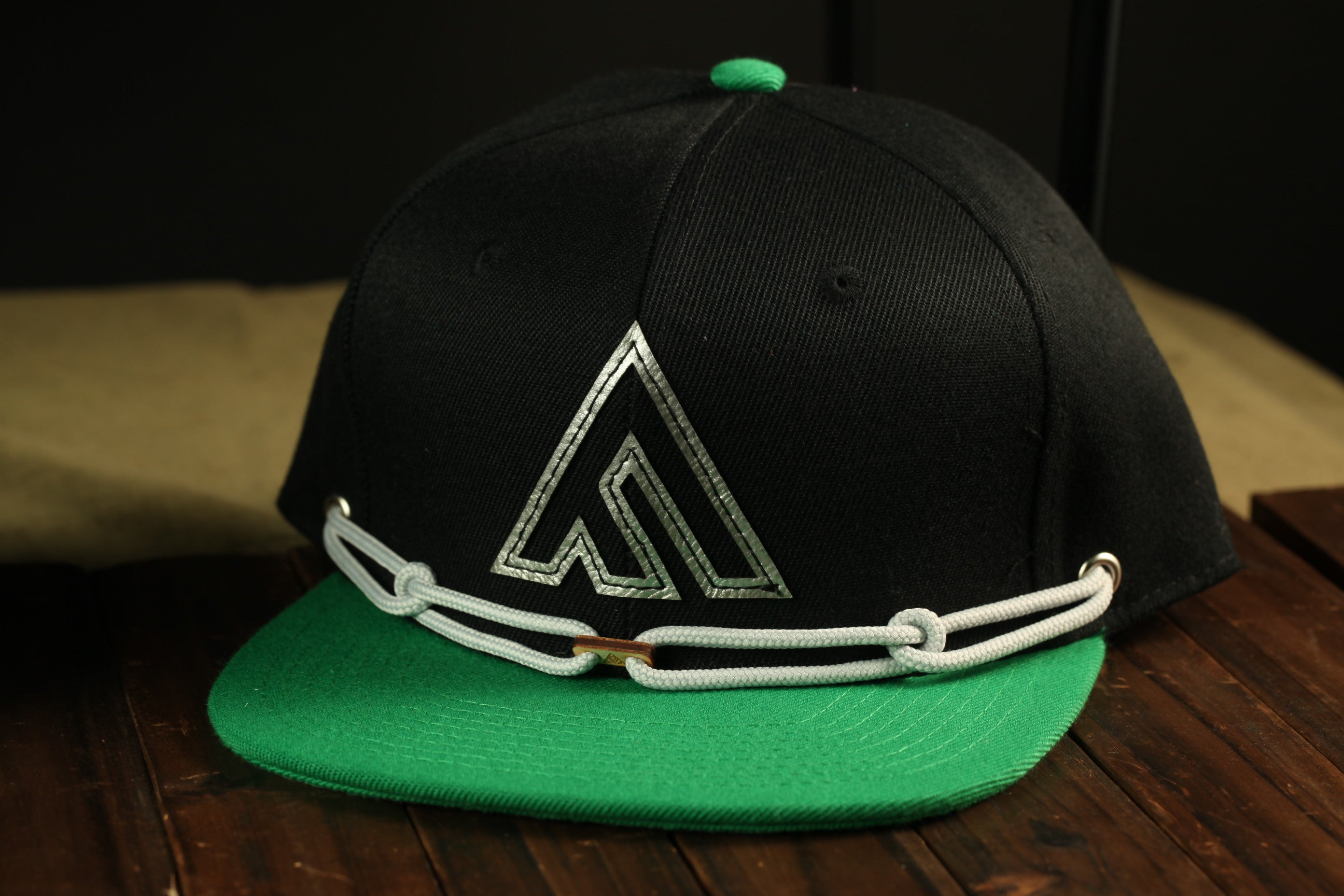 A black baseball cap with a green brim and a white logo on the front, sitting on a wooden surface.