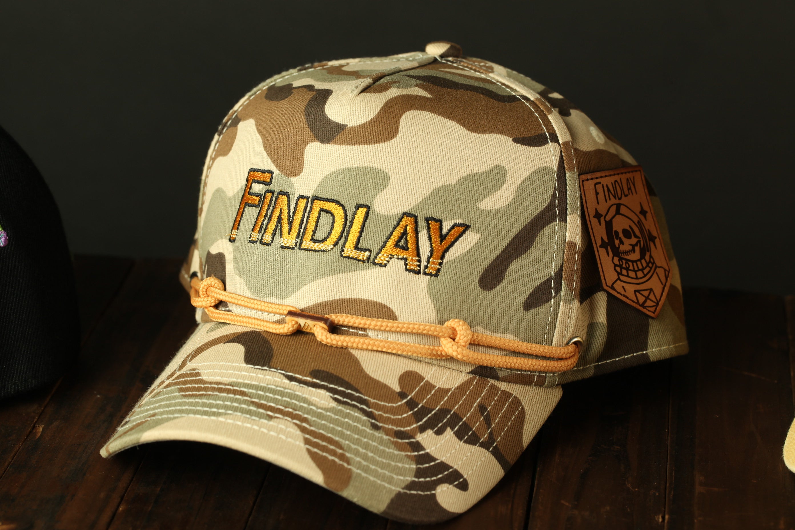 A camouflage baseball cap with the word "Findlay" printed on it, sitting on a wooden surface.