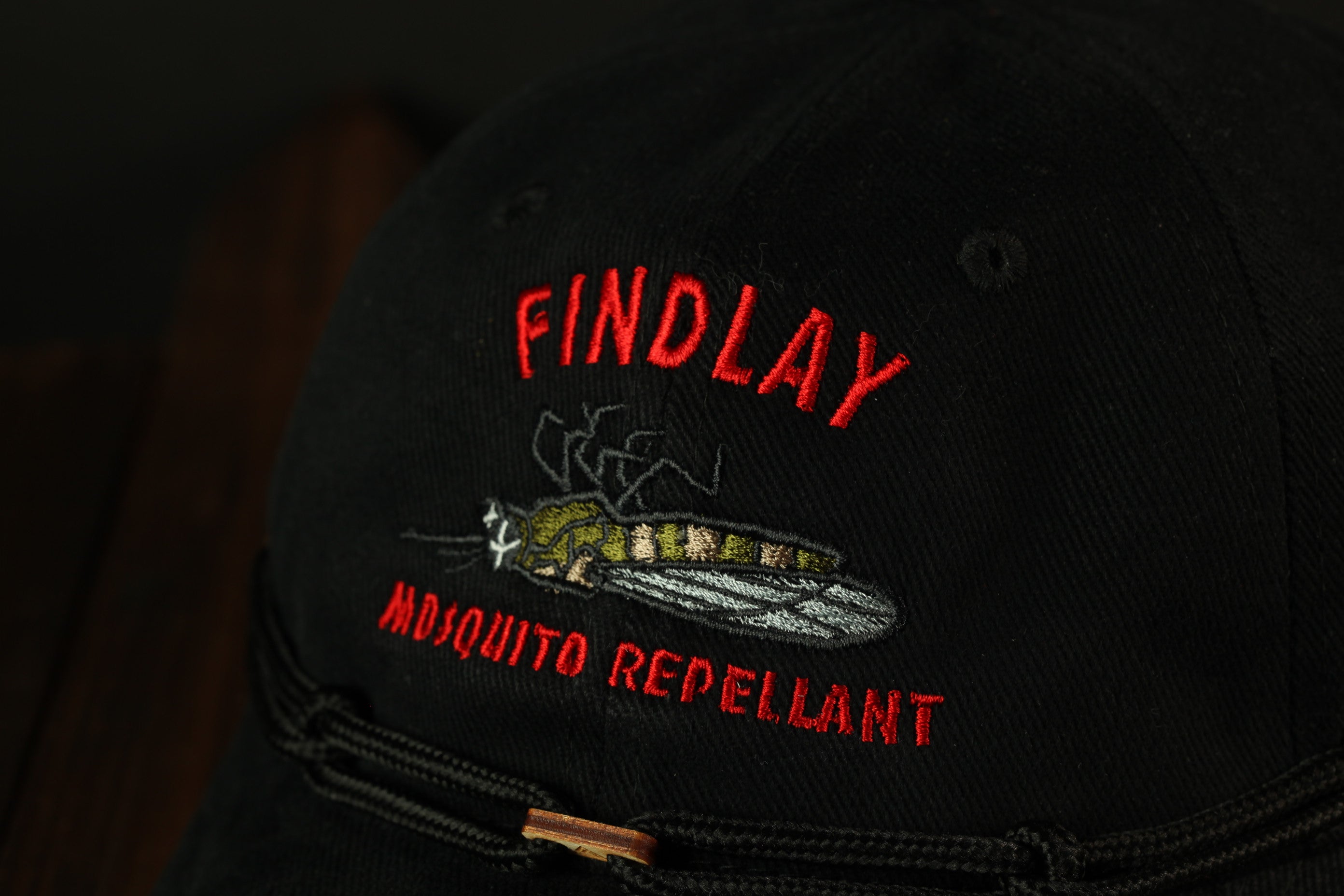A close-up view of a black baseball cap with red embroidered text and an image of a mosquito.