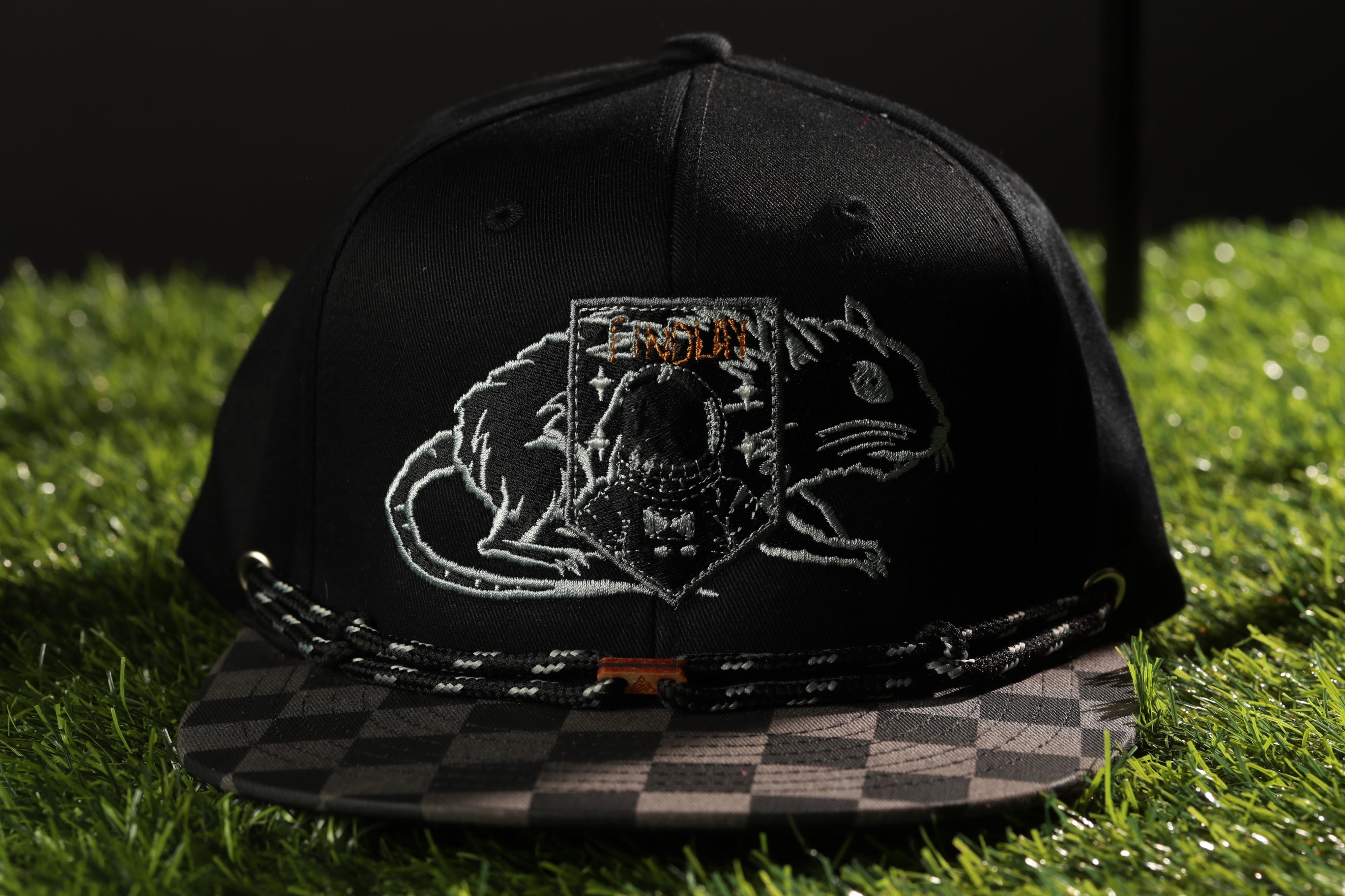 A black baseball cap with a checkered pattern and a graphic of a cat on the front, placed on a grassy surface.