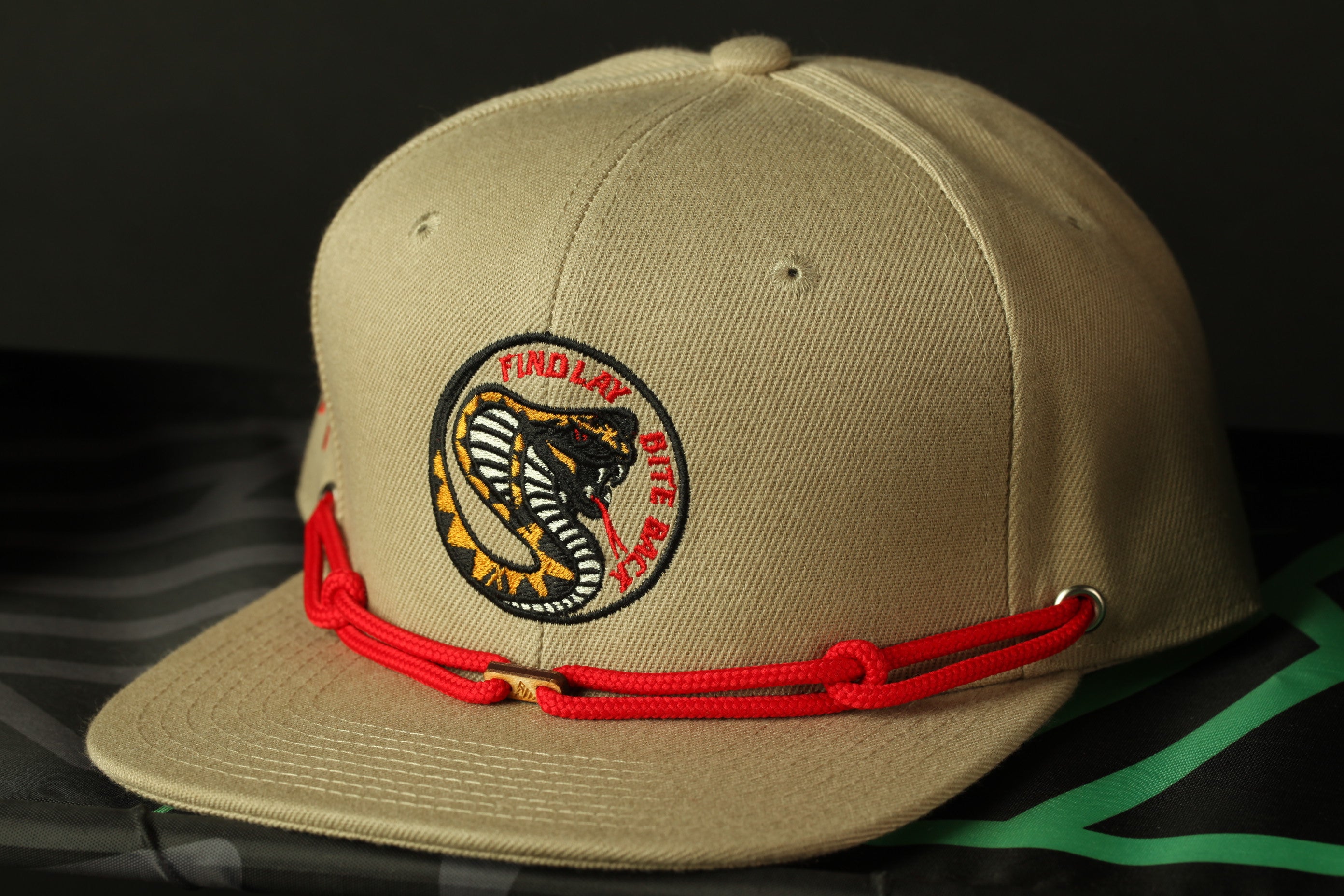 A beige baseball cap with a red string and a logo on the front, placed on a black and green striped surface.