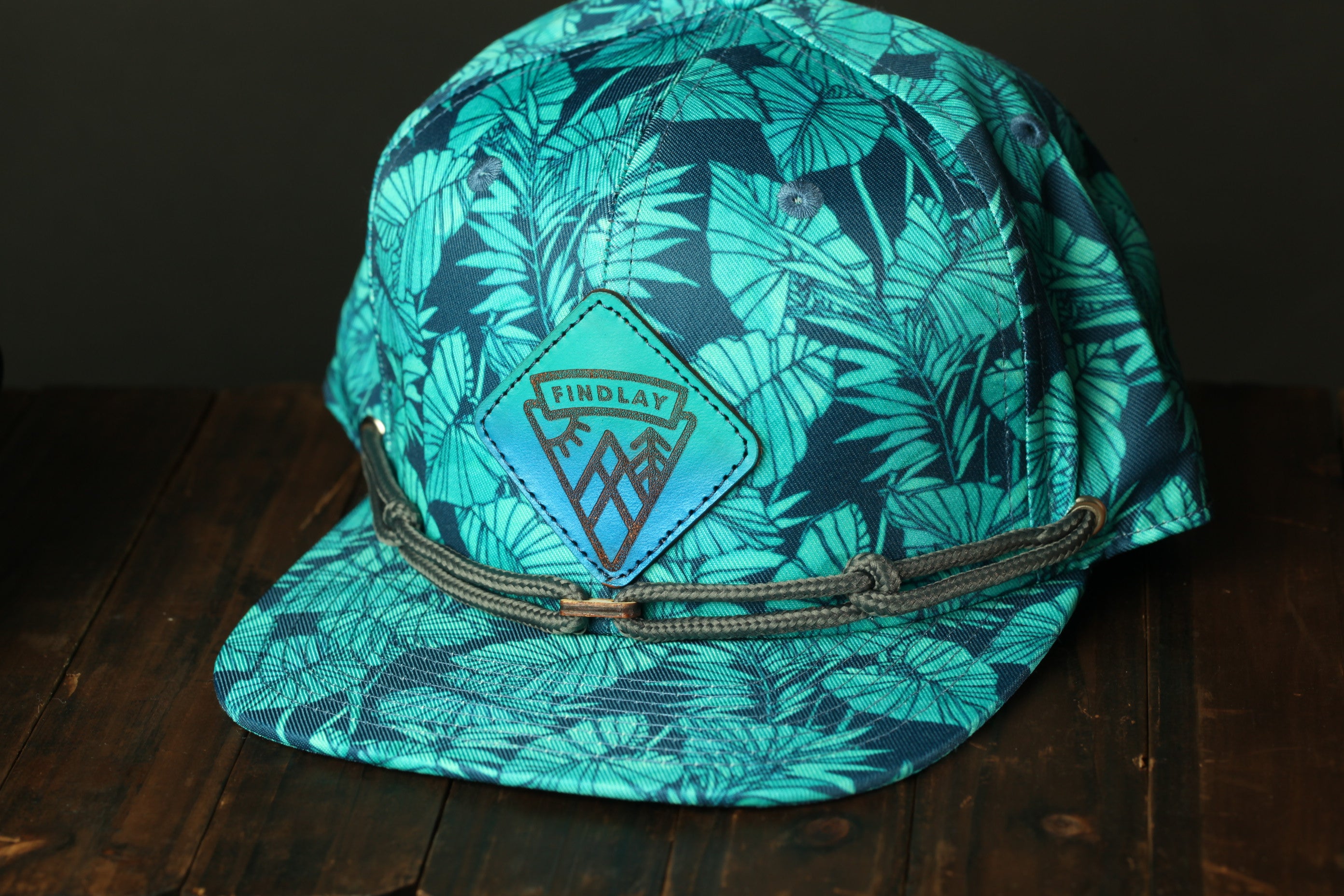 A turquoise baseball cap with a leaf pattern and a "Findlay" logo on the front, placed on a wooden surface.