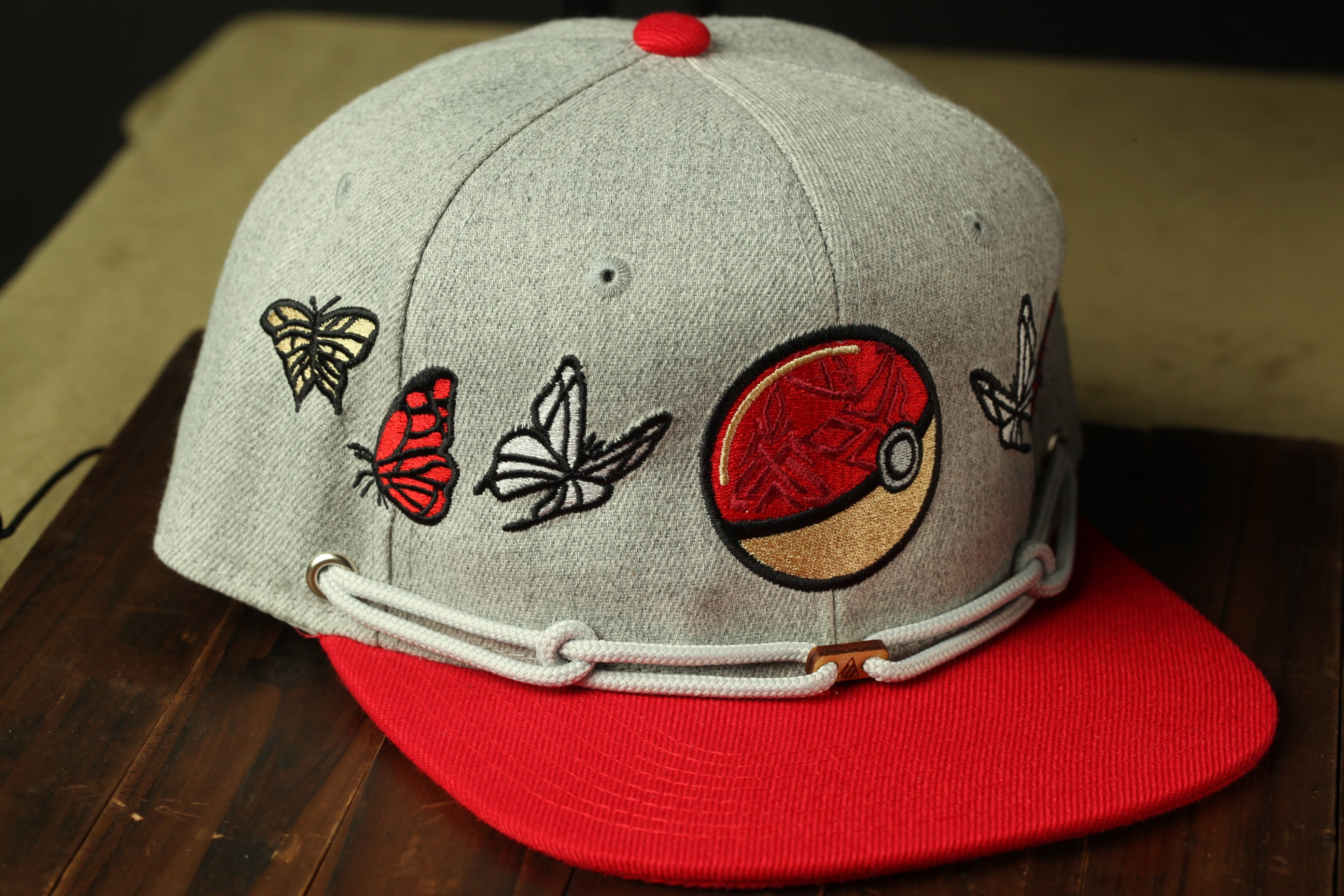 A gray baseball cap with a red brim, adorned with various embroidered designs, including butterflies and a Pokemon ball.
