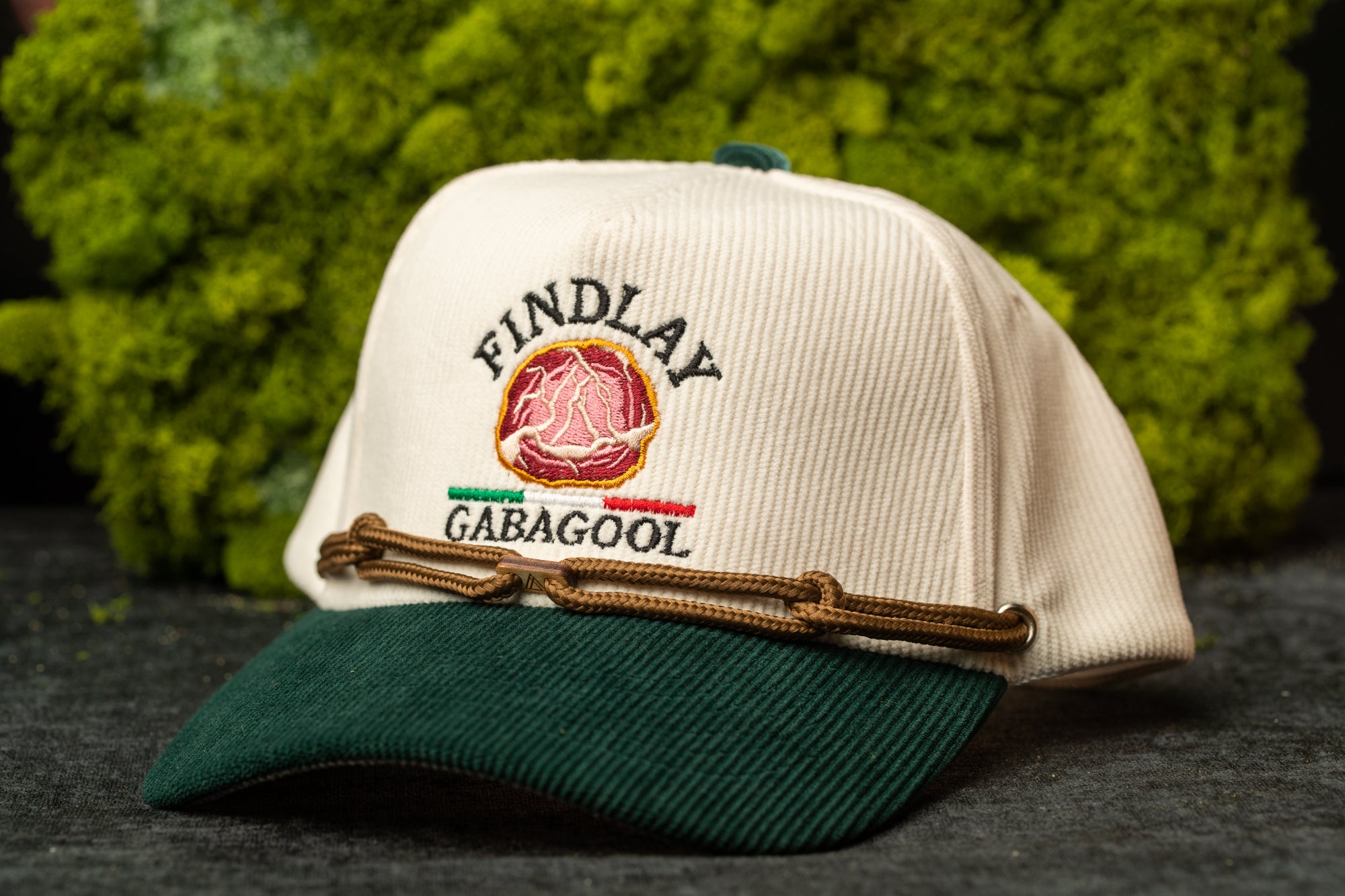A white baseball cap with a green brim and a brown leather strap, featuring embroidered text and a logo, is displayed against a background of green moss.