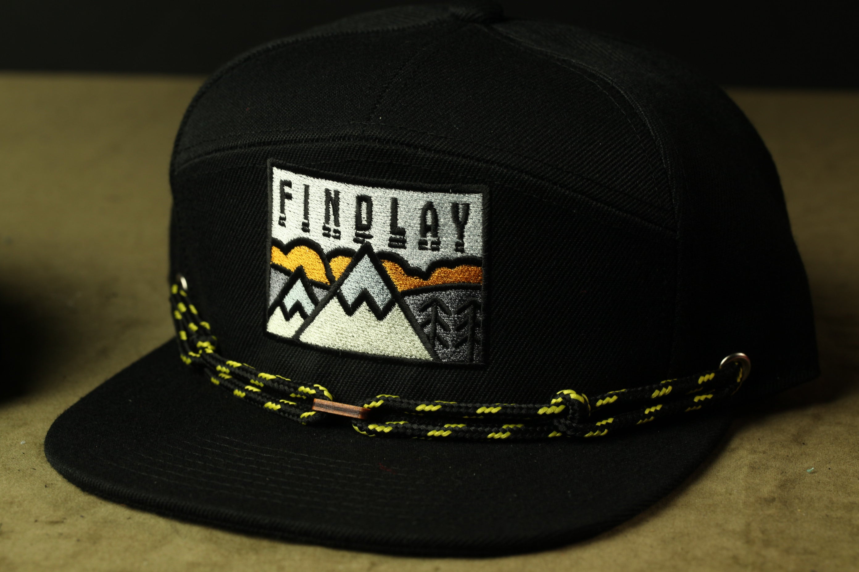 A black baseball cap with a yellow and white mountain design and the word "FINDOLAY" printed on it.