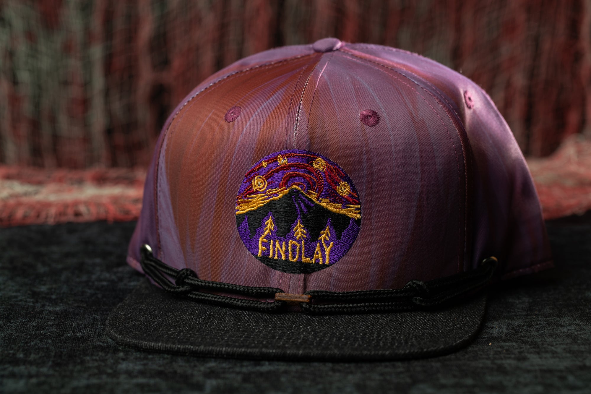 A purple baseball cap with a black brim and a circular embroidered logo on the front, featuring a mountain landscape and the word "FINCLAY".