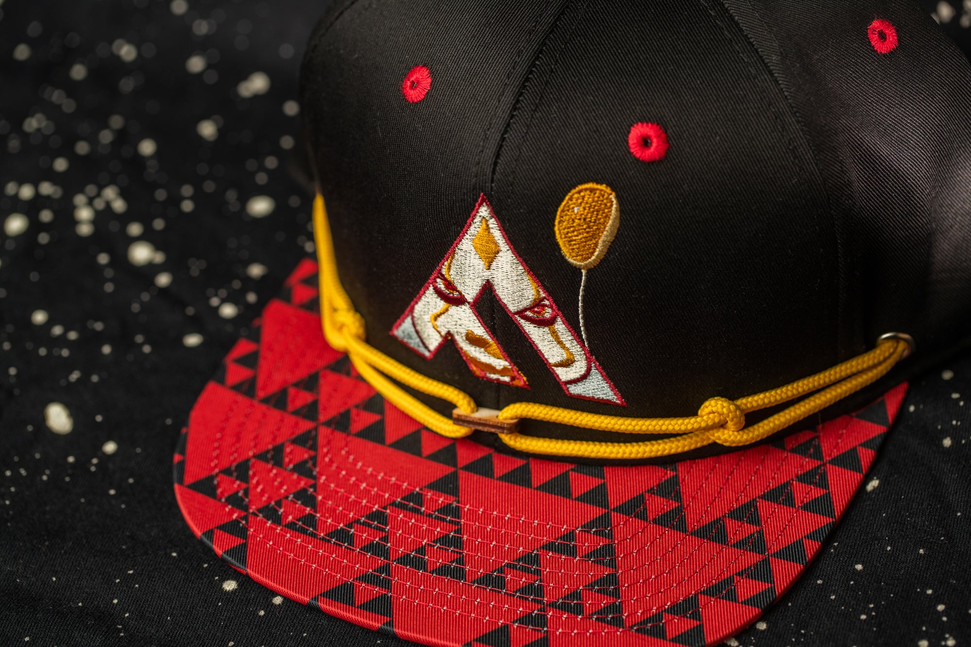 A black baseball cap with red and yellow accents, including a logo and a balloon design, is shown against a black background with a red and white geometric pattern.