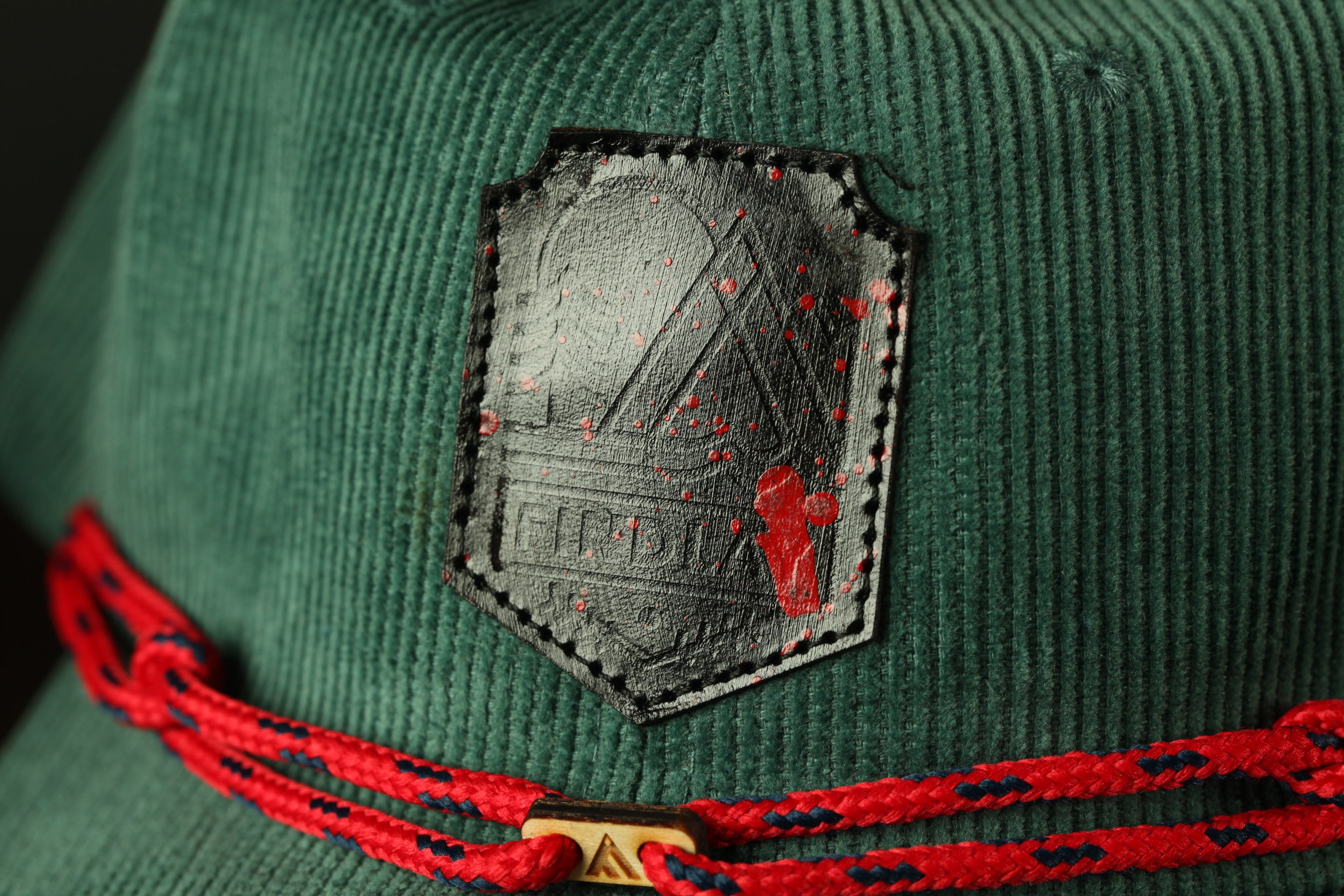 A close-up view of a green corduroy hat with a black patch featuring a red logo.