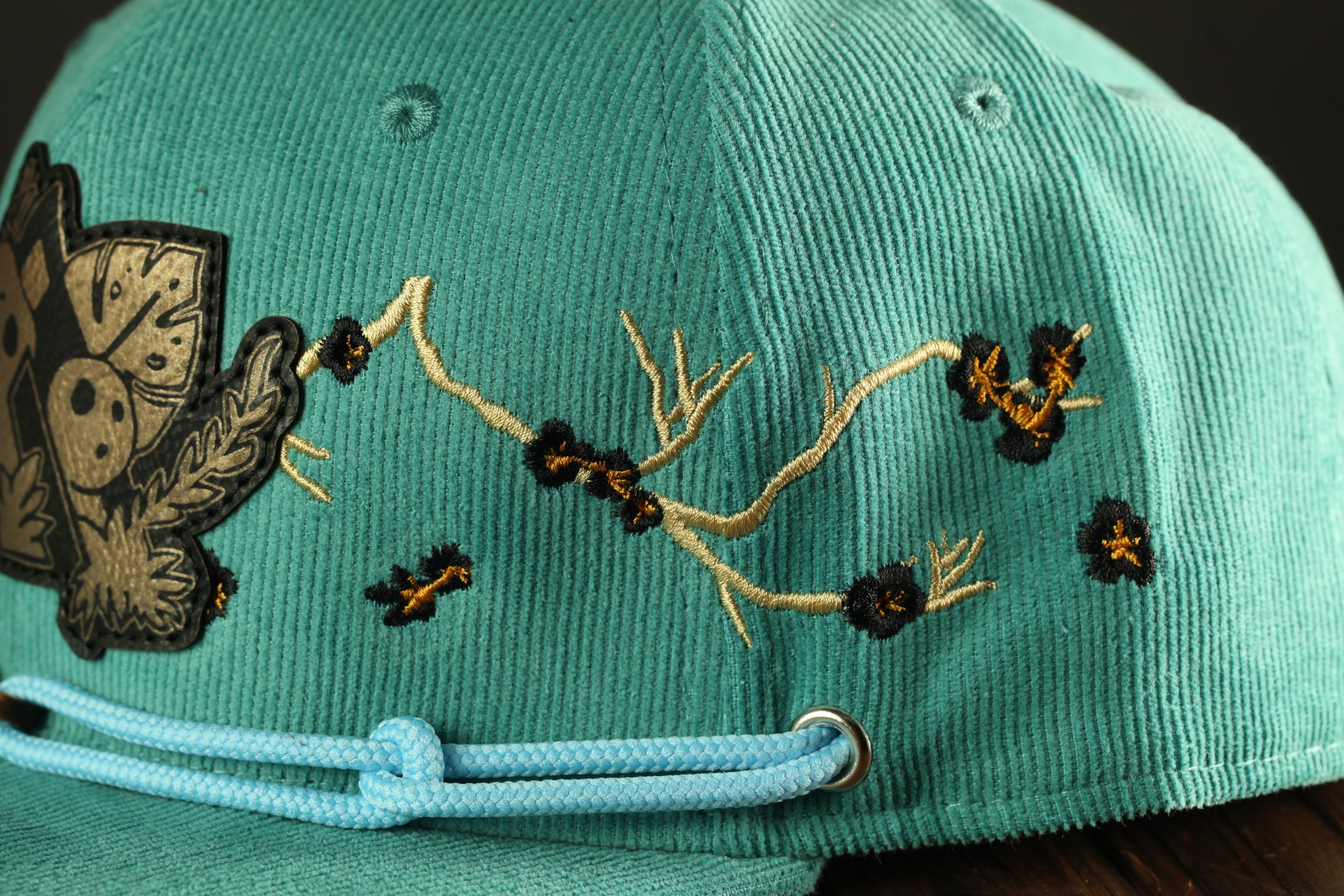 A close-up view of a turquoise baseball cap with intricate embroidered designs, including floral patterns and small black and gold accents.