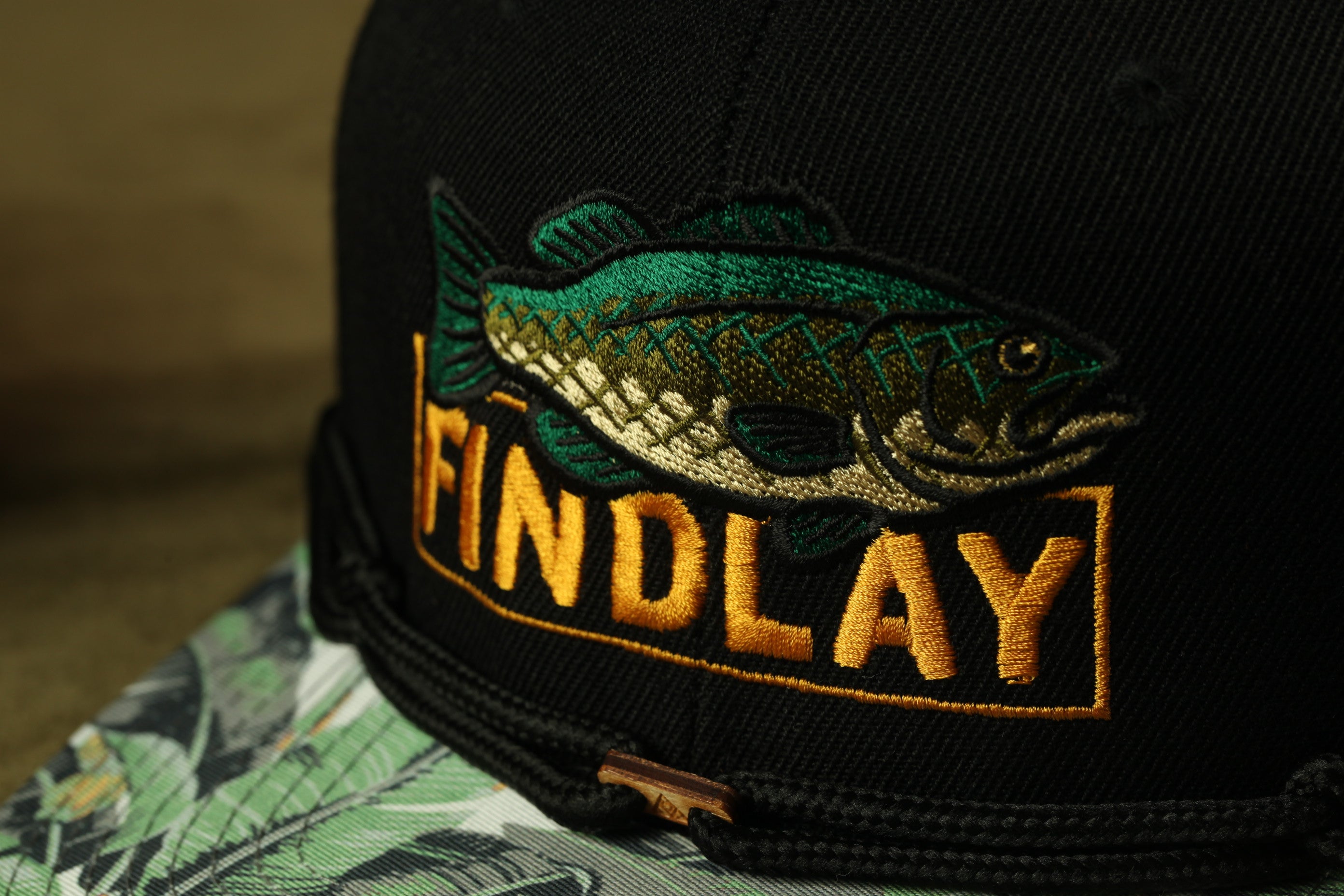 A close-up view of a black baseball cap with a fish embroidered on it, featuring the word "FINDLAY" in gold letters.