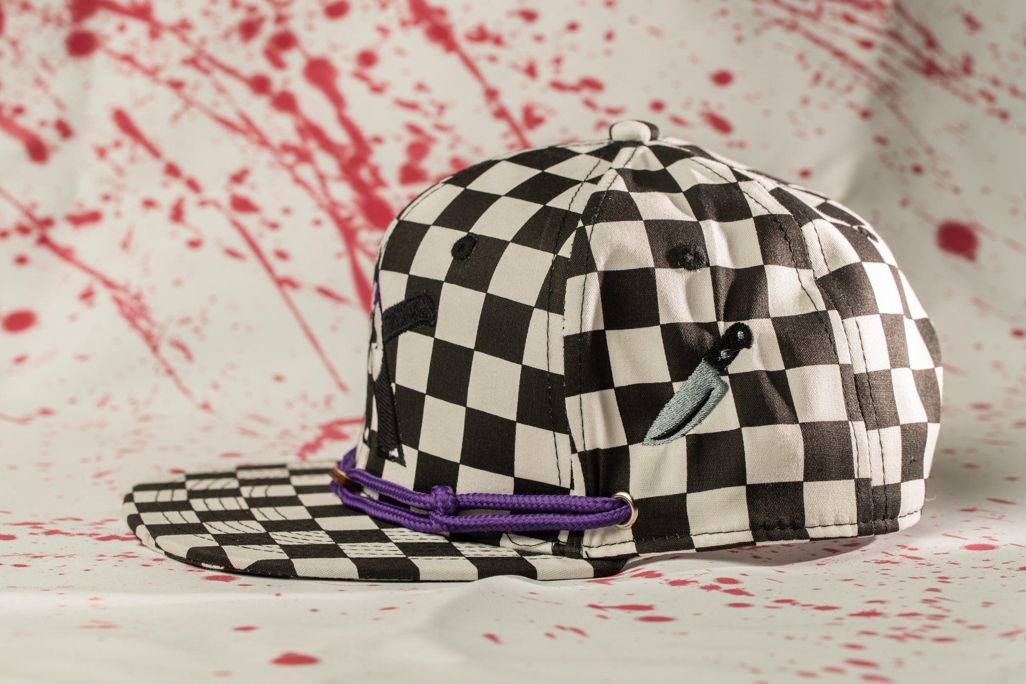 A black and white checkered baseball cap with a purple string and a small knife-like object attached to it, set against a background with red splatters.