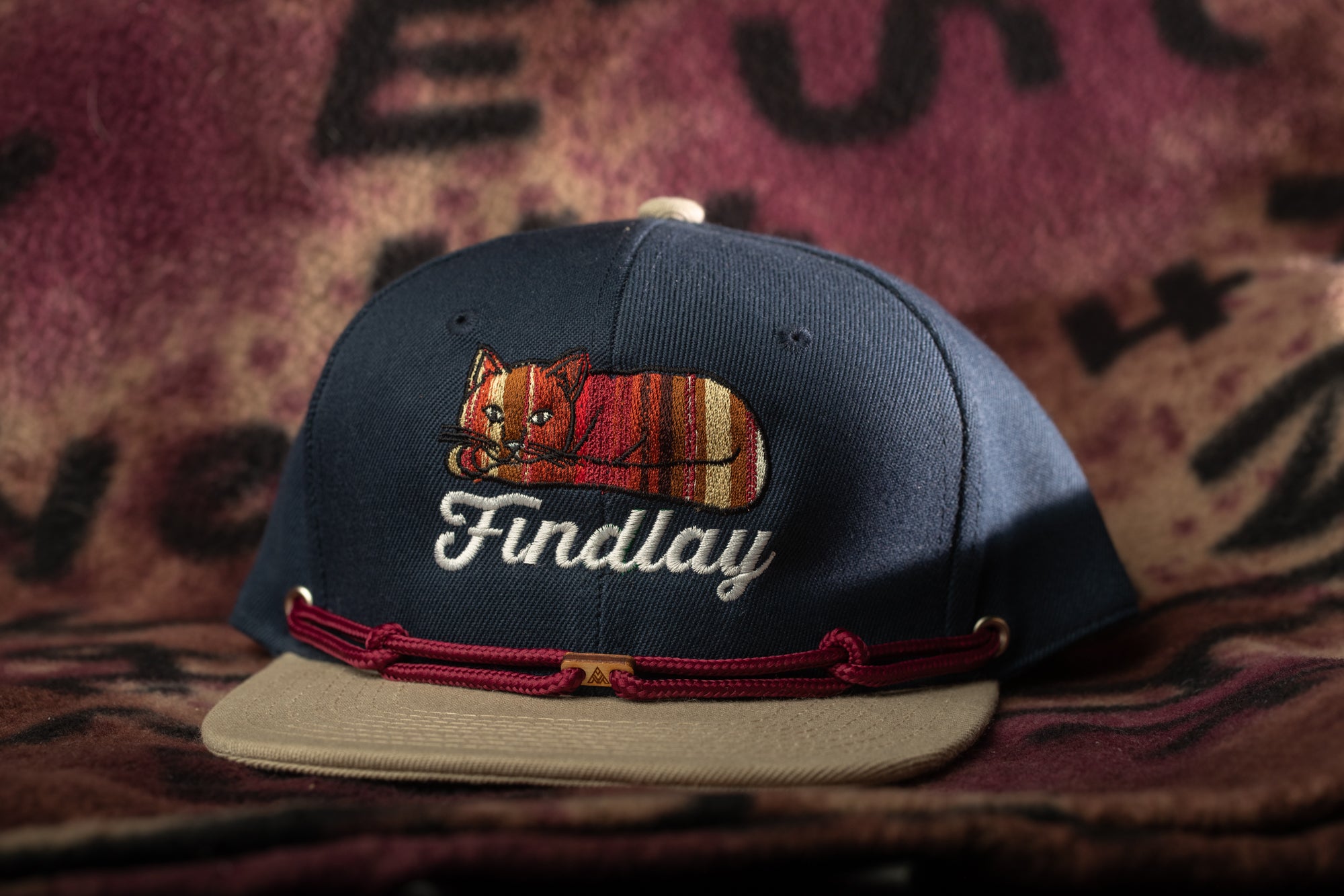 A navy blue baseball cap with a cat embroidered on it, sitting on a beige fabric surface.
