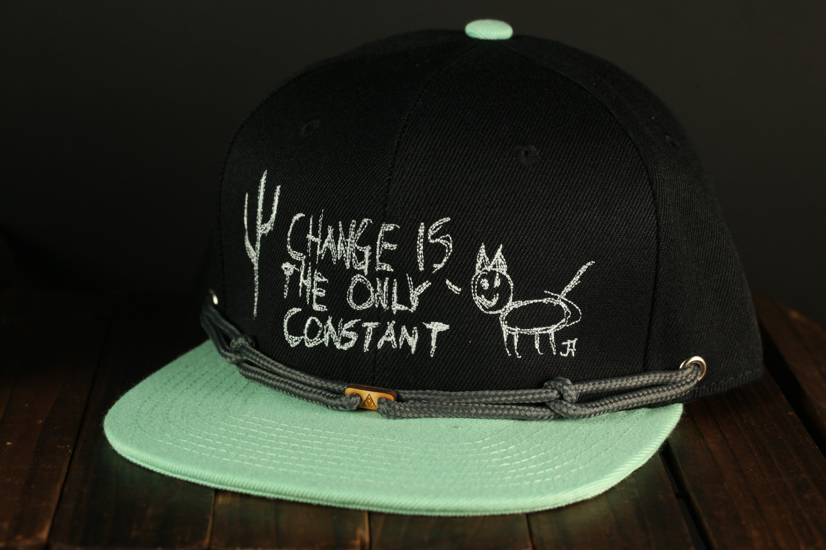 A black baseball cap with a green brim, featuring white text and a drawing of a cat.