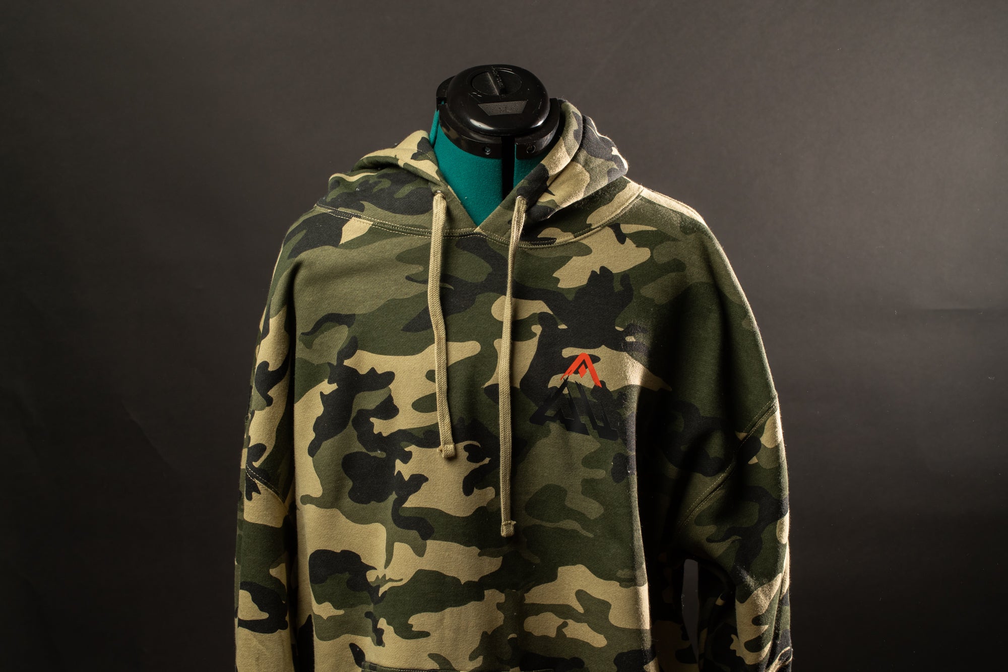 A camouflage hoodie with a red logo on the left side is displayed against a dark background.