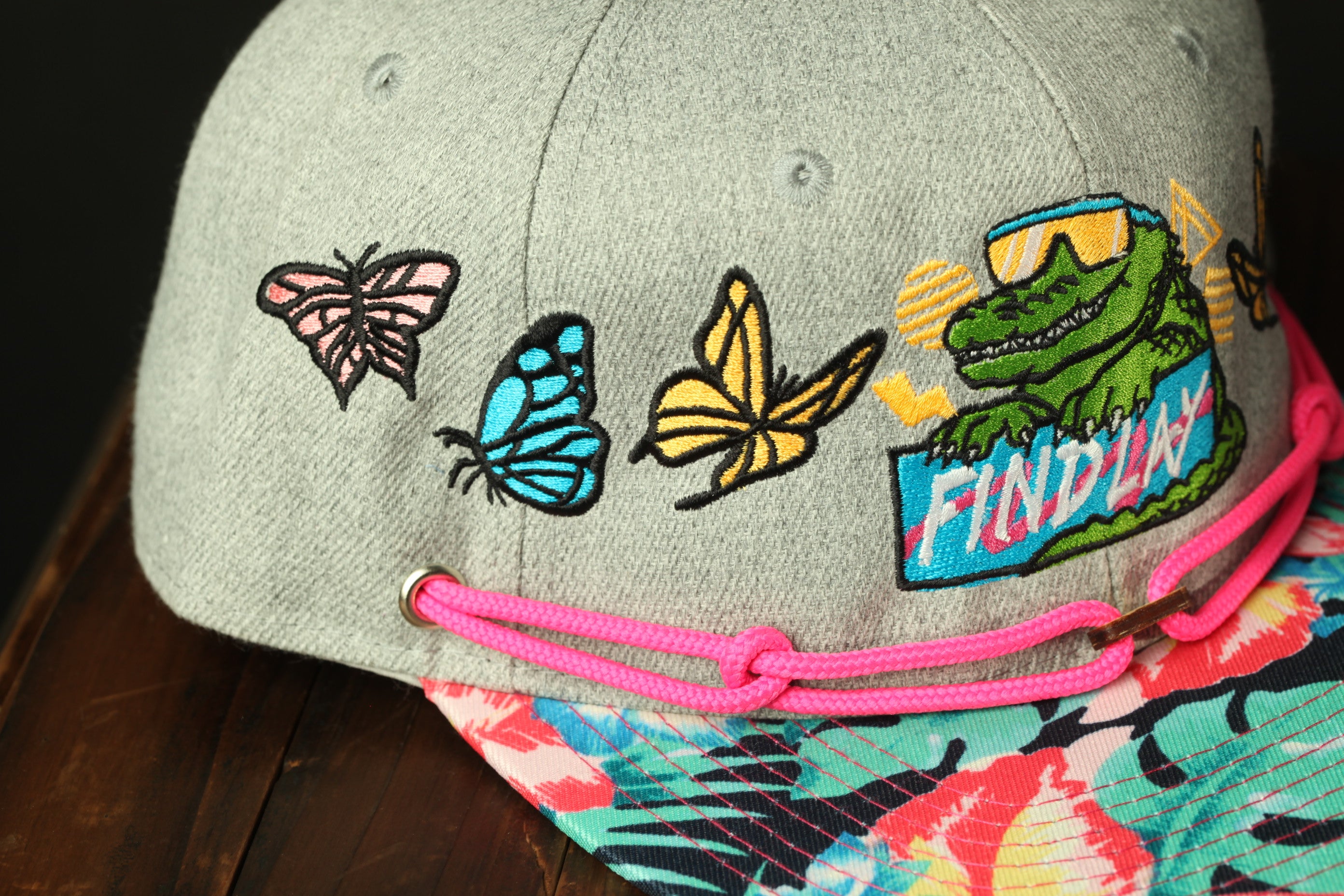 A gray baseball cap with colorful embroidered designs, including a green dinosaur, butterflies, and a pink string.