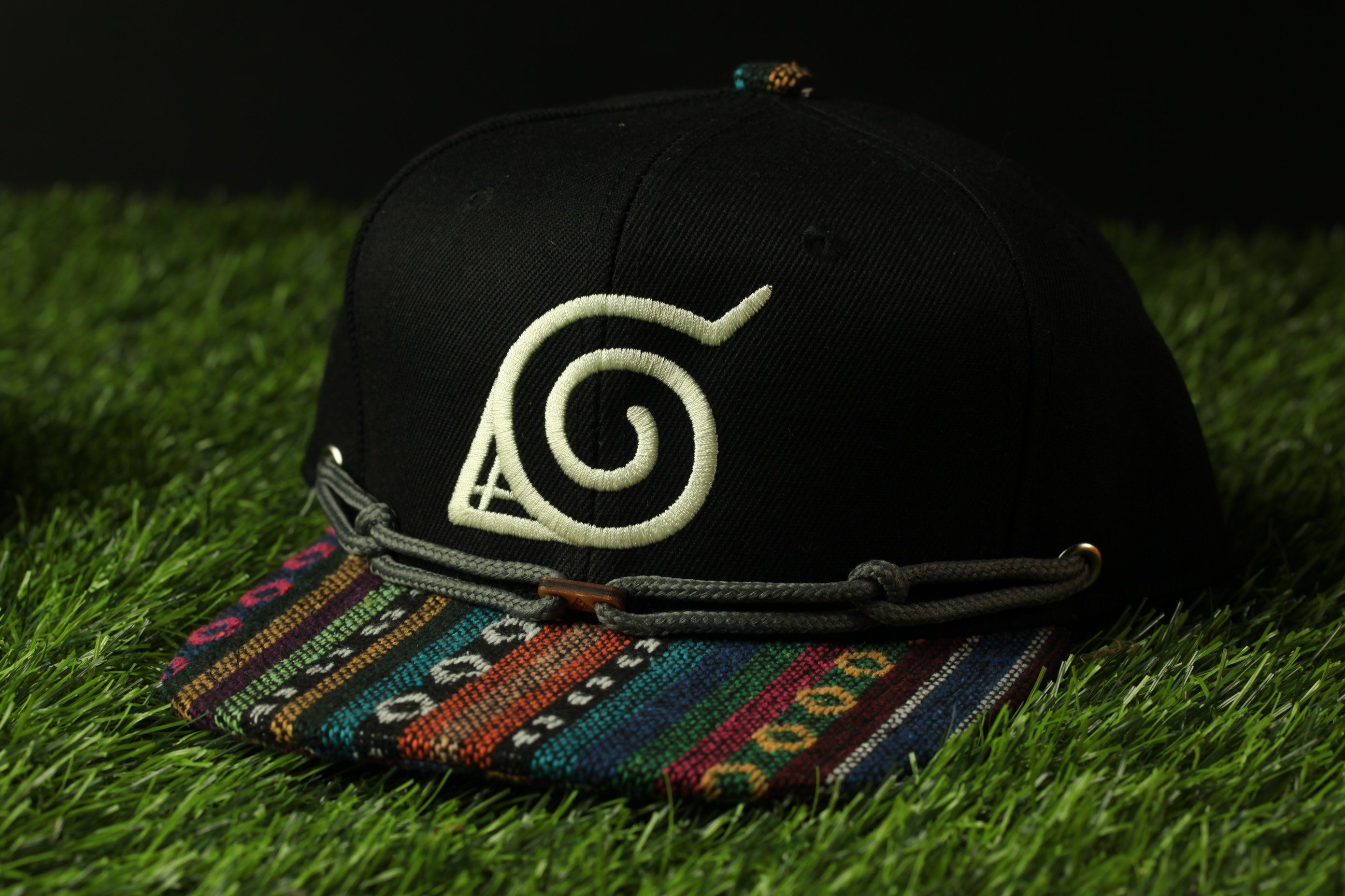 A black baseball cap with a colorful, patterned brim and a white, stylized logo on the front, resting on a grassy surface.