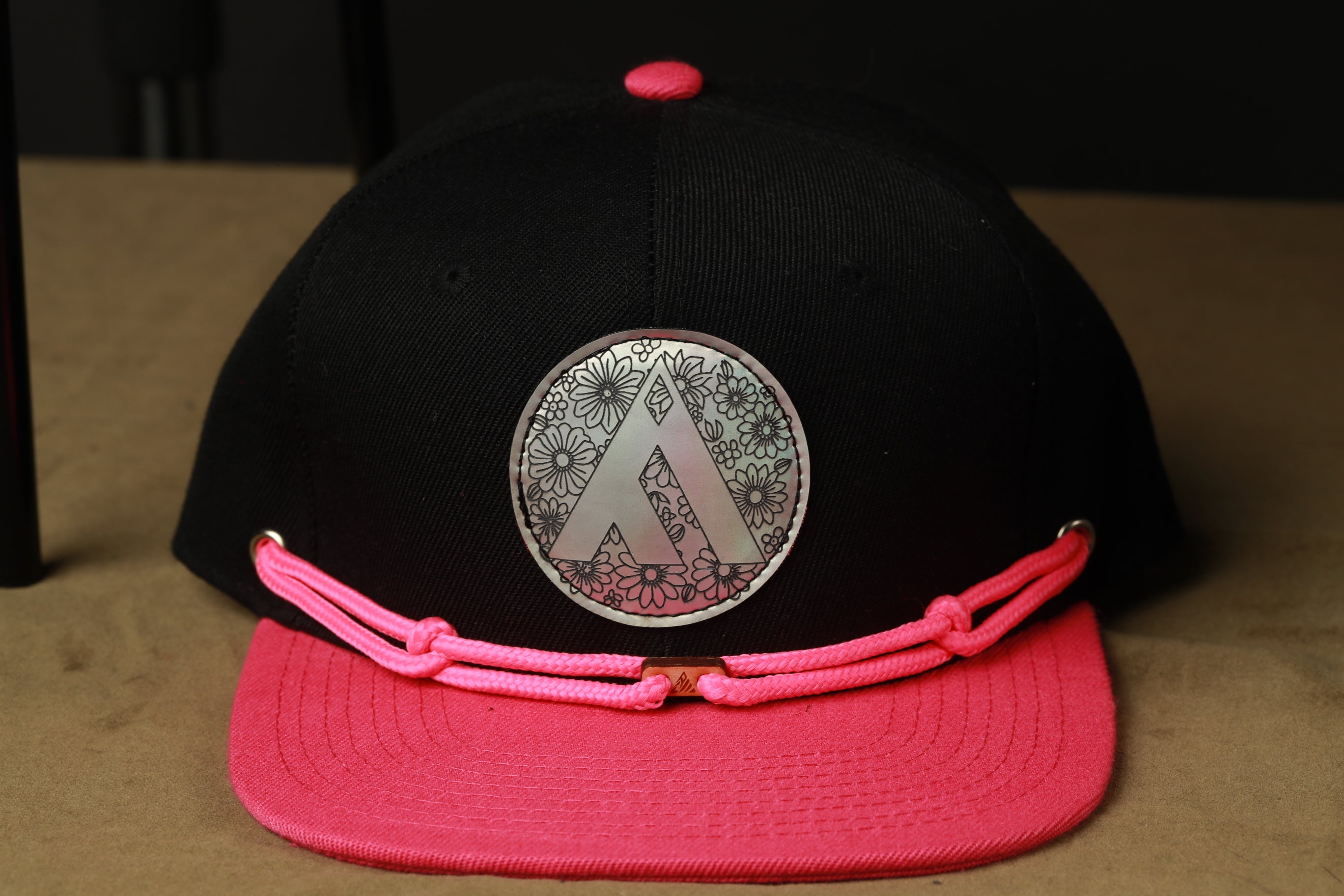A black baseball cap with a pink brim and a silver circular logo on the front.