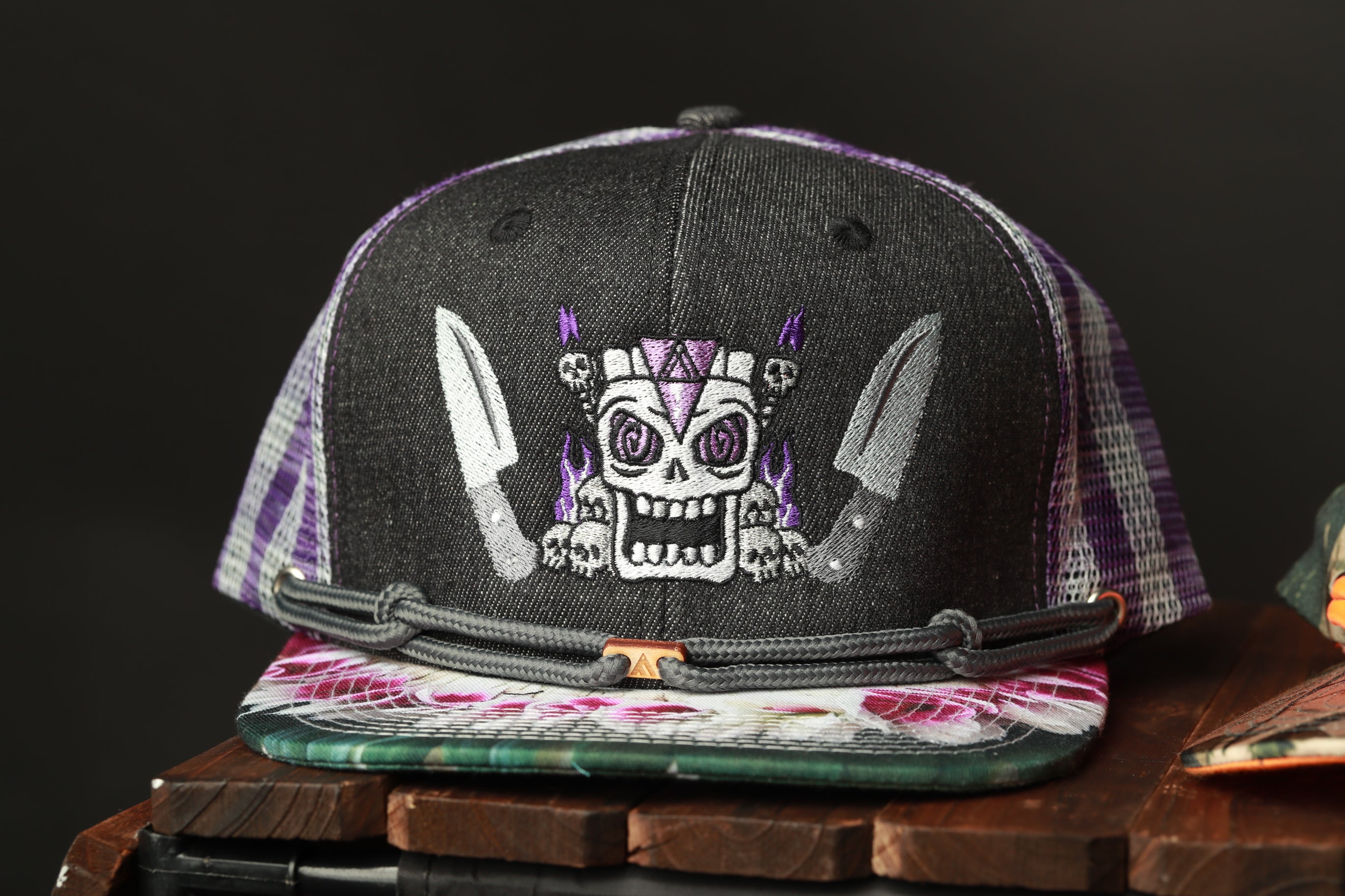 A black baseball cap with a skull and crossbones design, featuring purple and white plaid trim, is displayed on a wooden surface.
