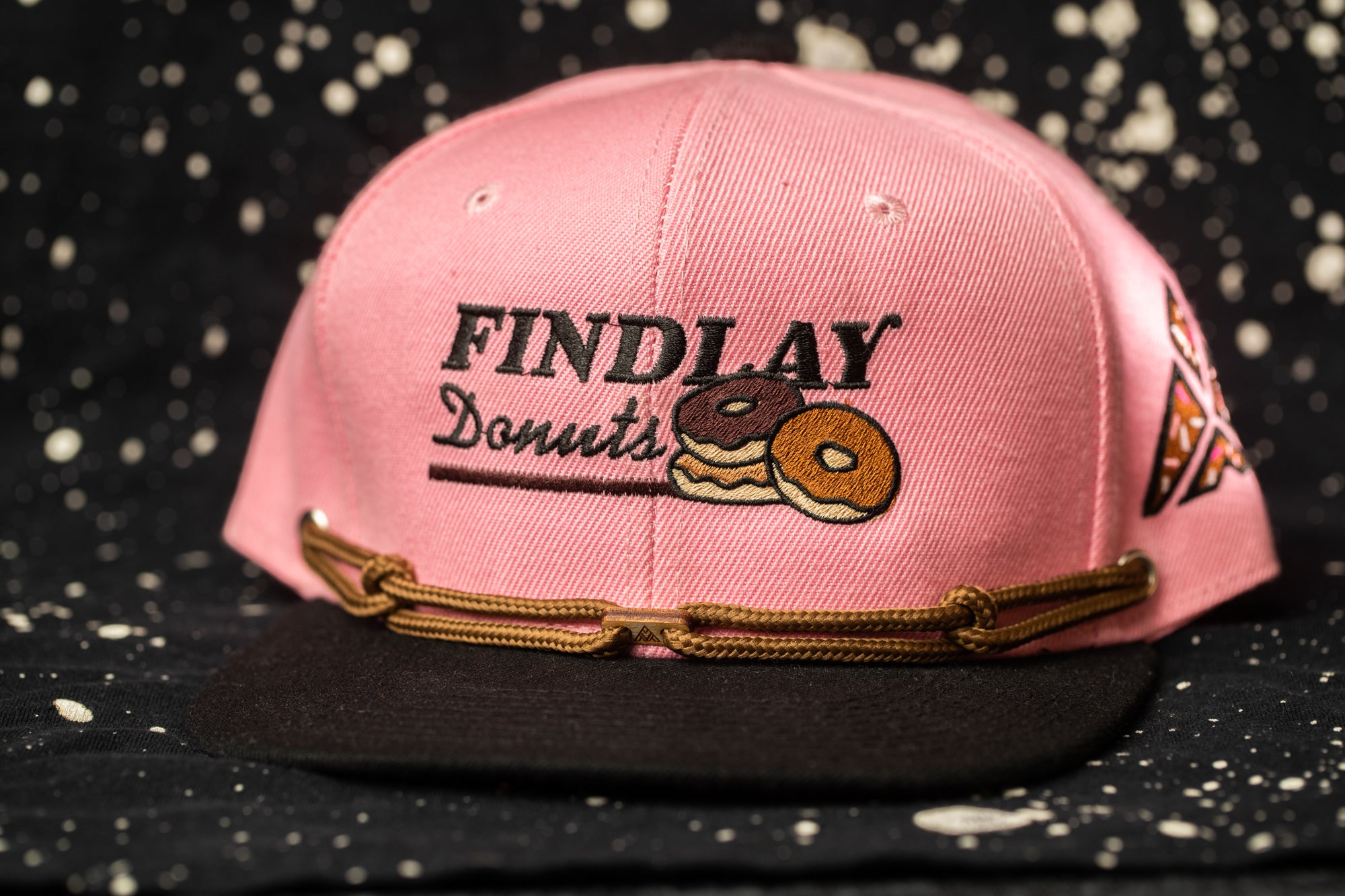 A pink baseball cap with the text "FINDlay Donuts" printed on it, along with an illustration of two donuts.