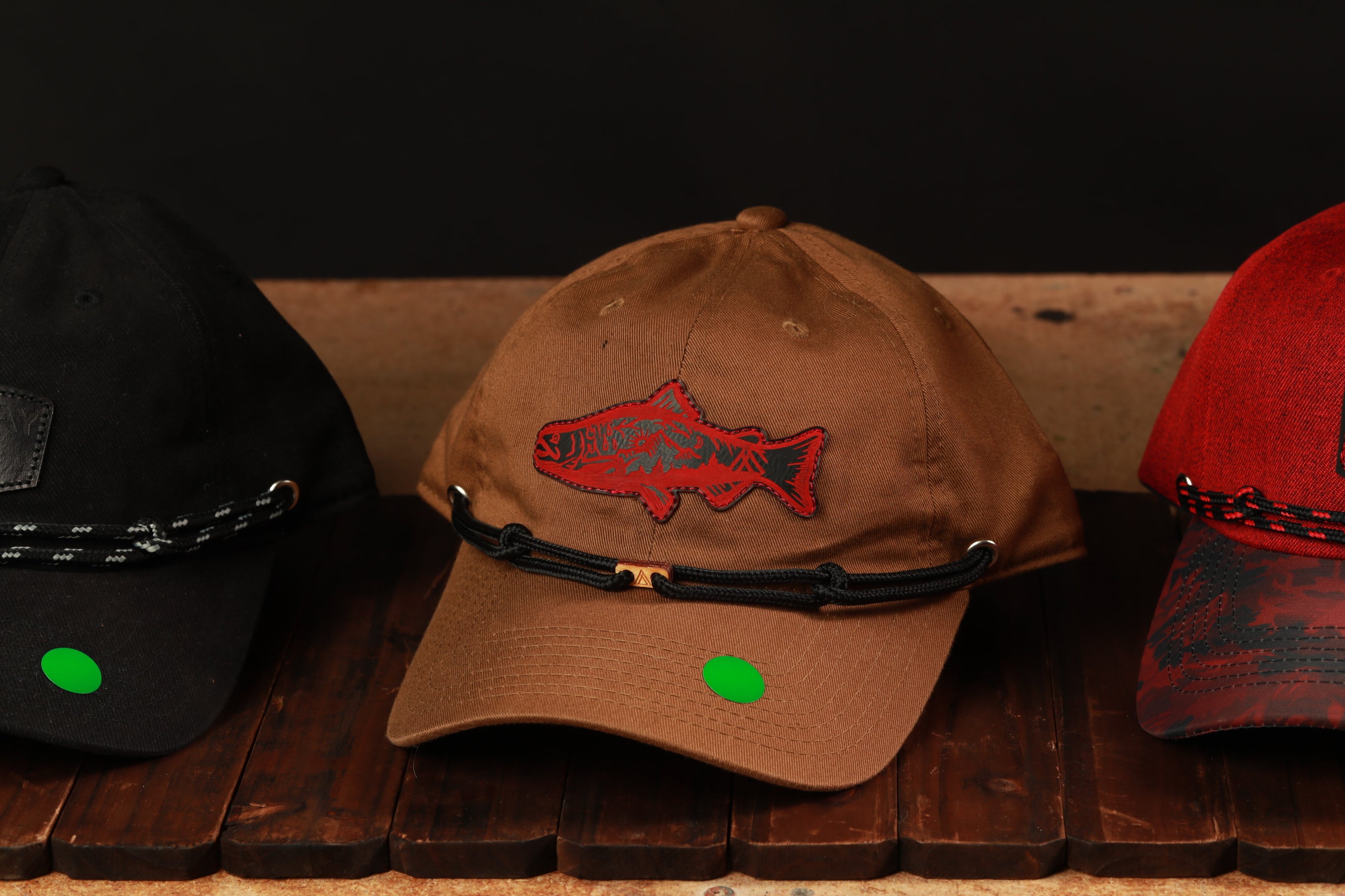 Three baseball caps are displayed on a wooden surface, with one featuring a fish embroidered on the front.