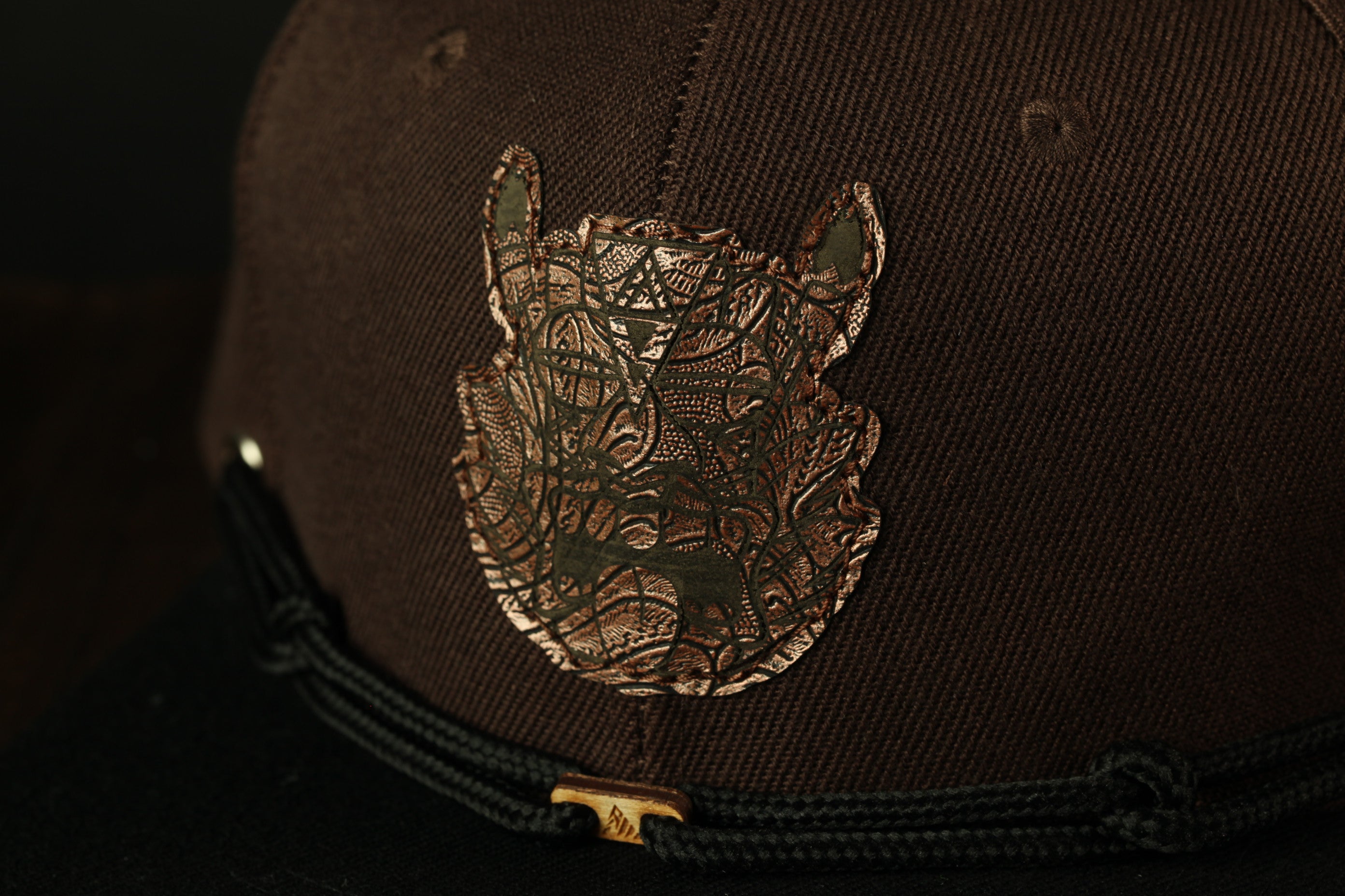 A close-up view of a dark brown baseball cap with a detailed embroidered design on the front.
