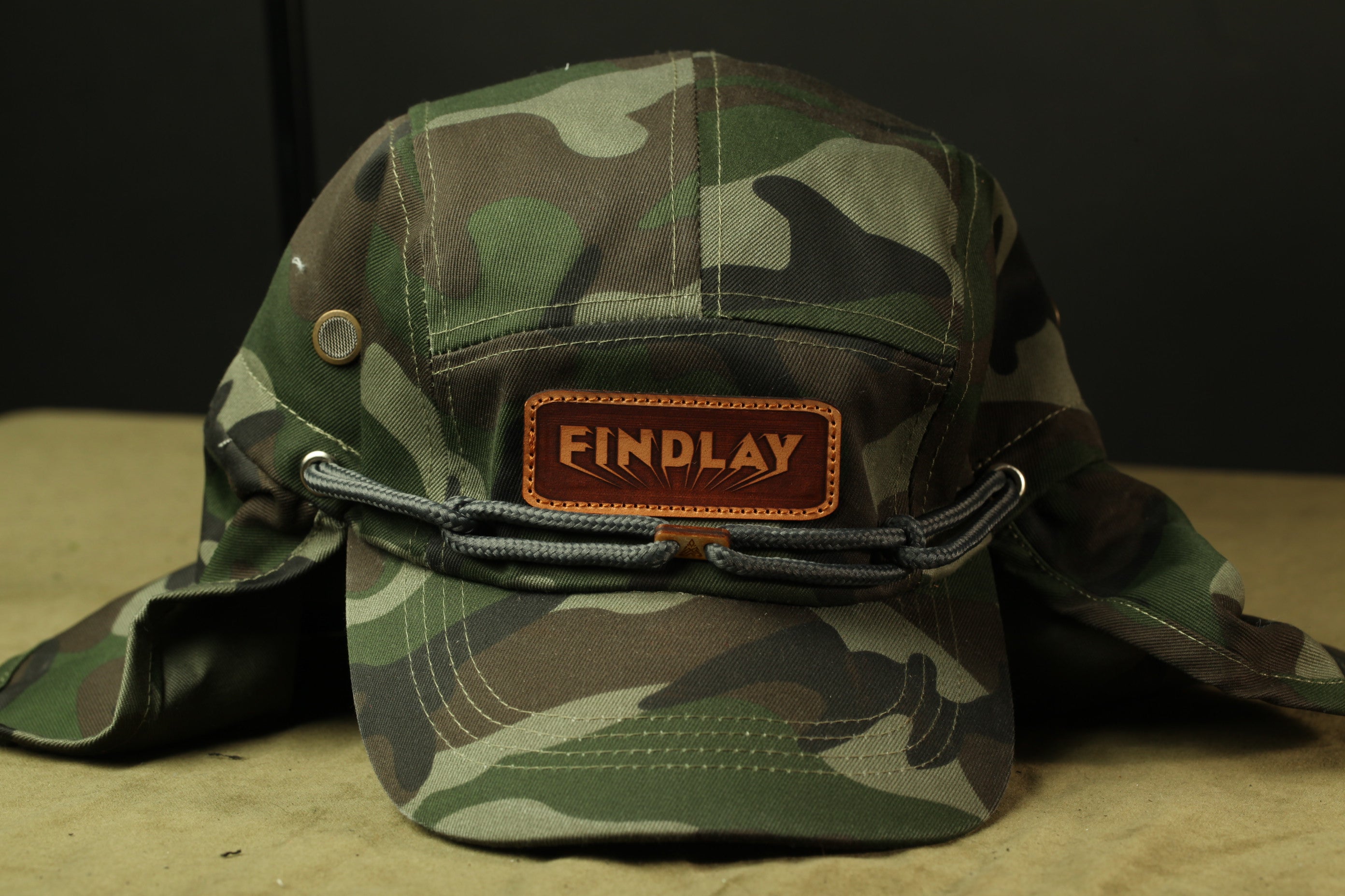 A camouflage-patterned military-style hat with a leather patch that says "FINDLAY" on it.